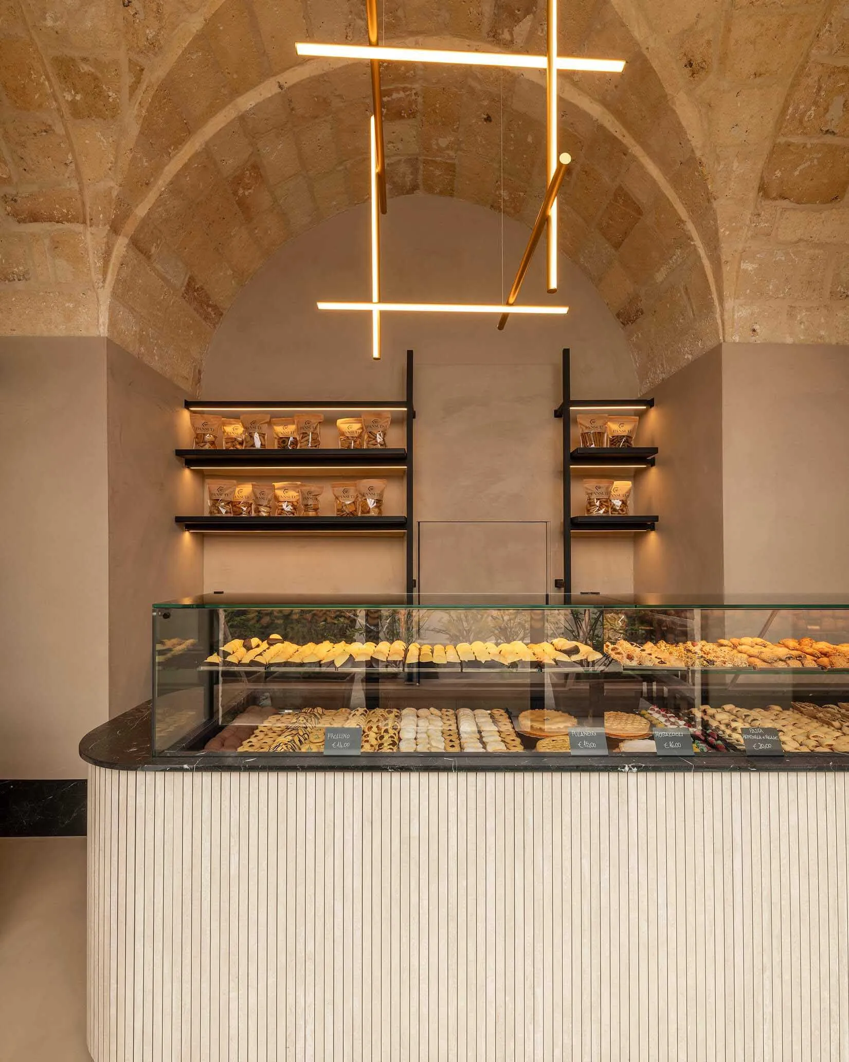 Modern bakery counter with glass pastry display filled with croissants and pastries, set against historic stone vaulted ceiling and illuminated floating shelves.