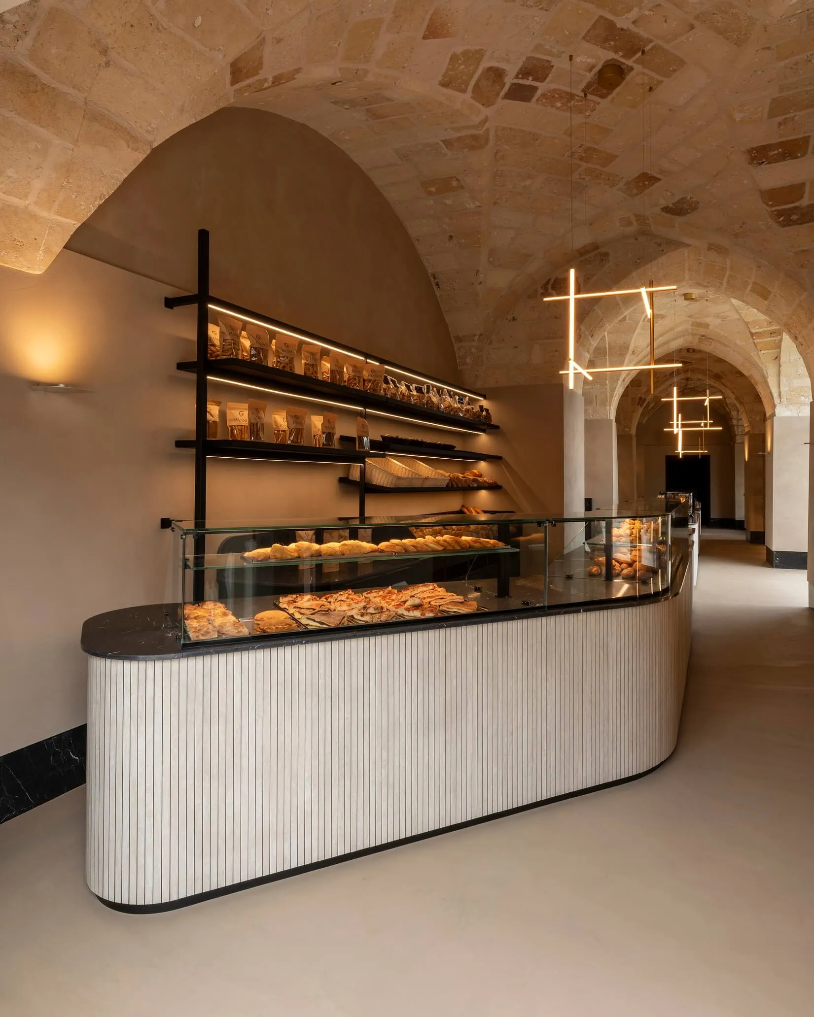 Modern bakery counter with curved fluted white paneling and black marble top, glass pastry display filled with breads and pastries, set against a historic stone vaulted ceiling with contemporary linear pendant lights.