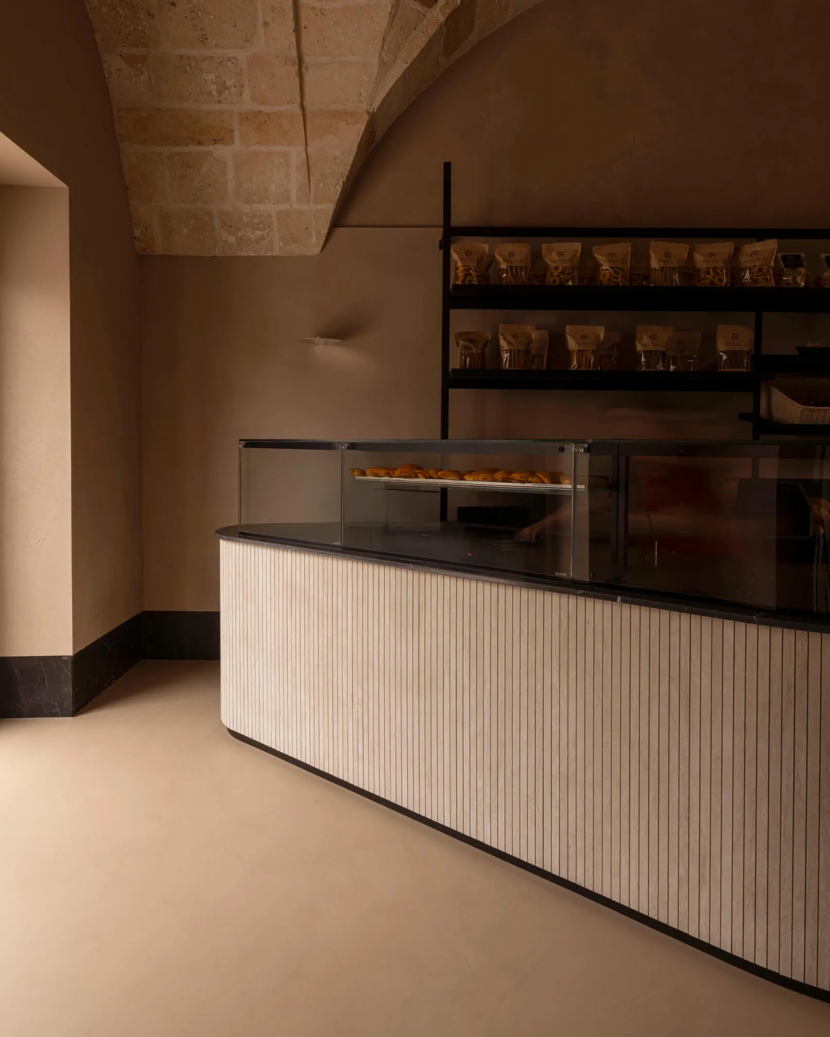 Modern curved bakery counter with vertical fluted wood paneling, glass pastry display case, and black metal shelving against a backdrop of original stone vaulted ceiling.
