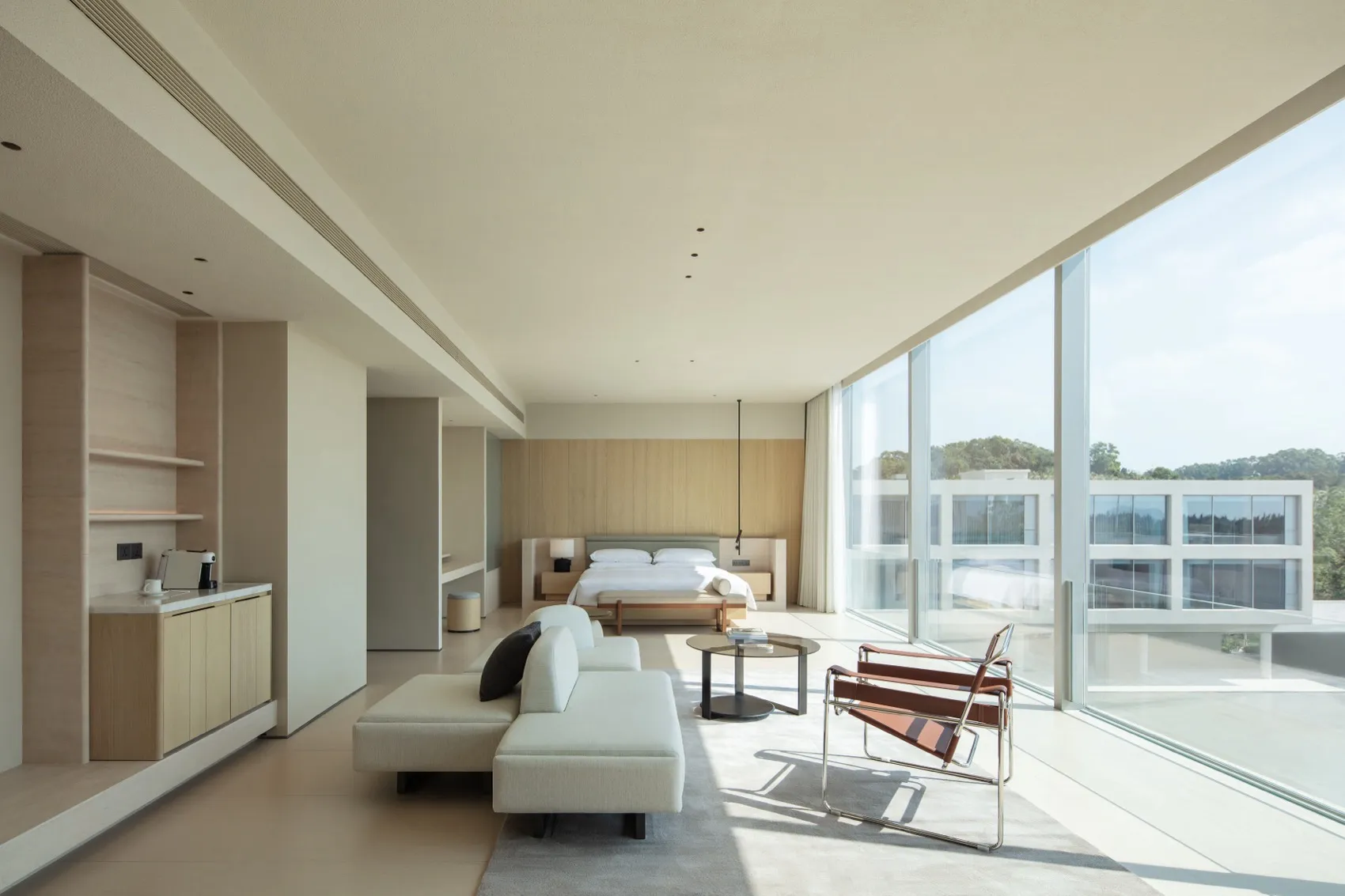 Modern minimalist bedroom with light oak cabinetry, white sectional sofa, brown leather sling chair, and floor-to-ceiling glass windows overlooking trees.