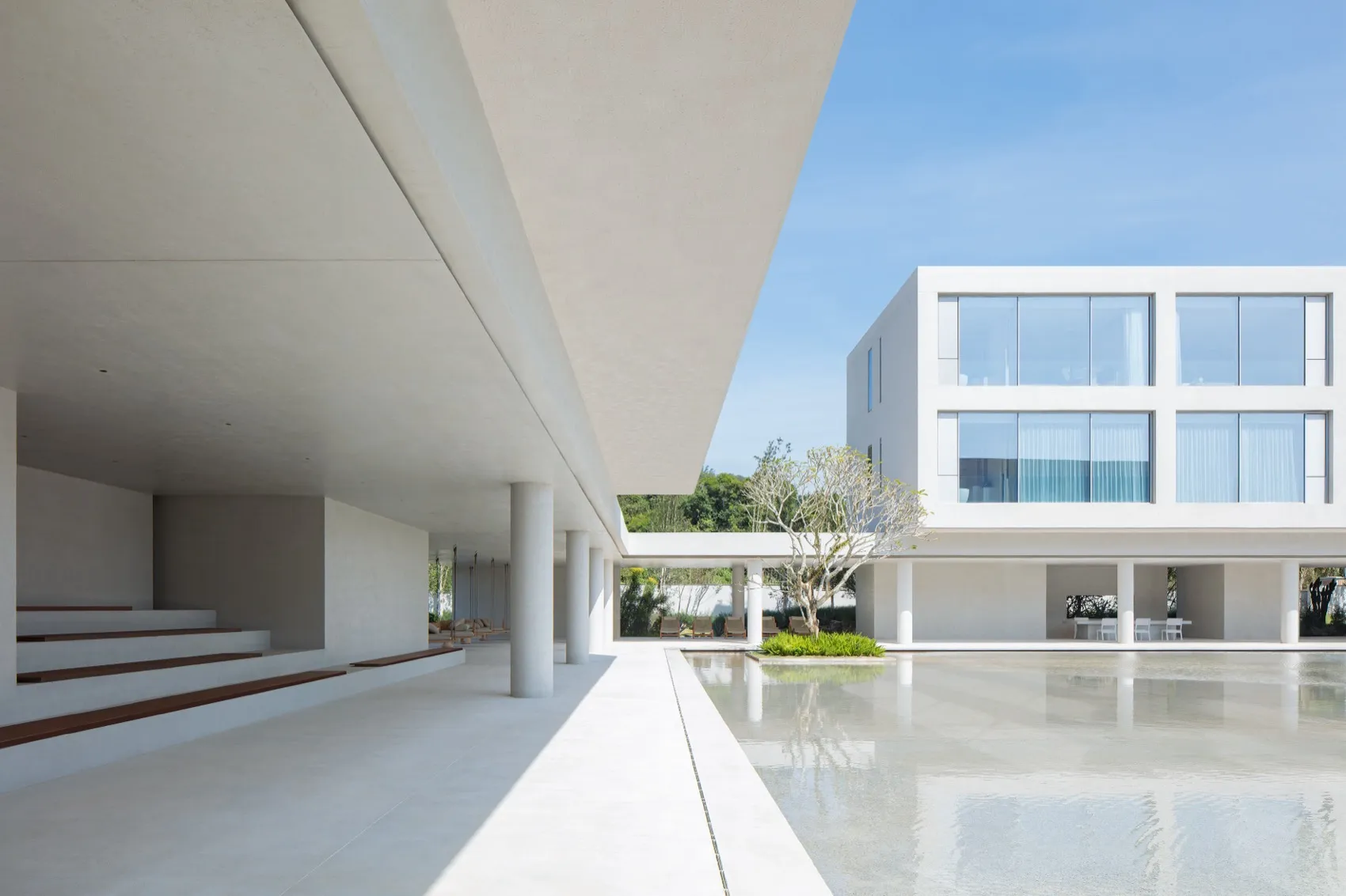 Modern minimalist concrete building with large reflecting pool in a sunlit courtyard featuring white pillars and a small tree.