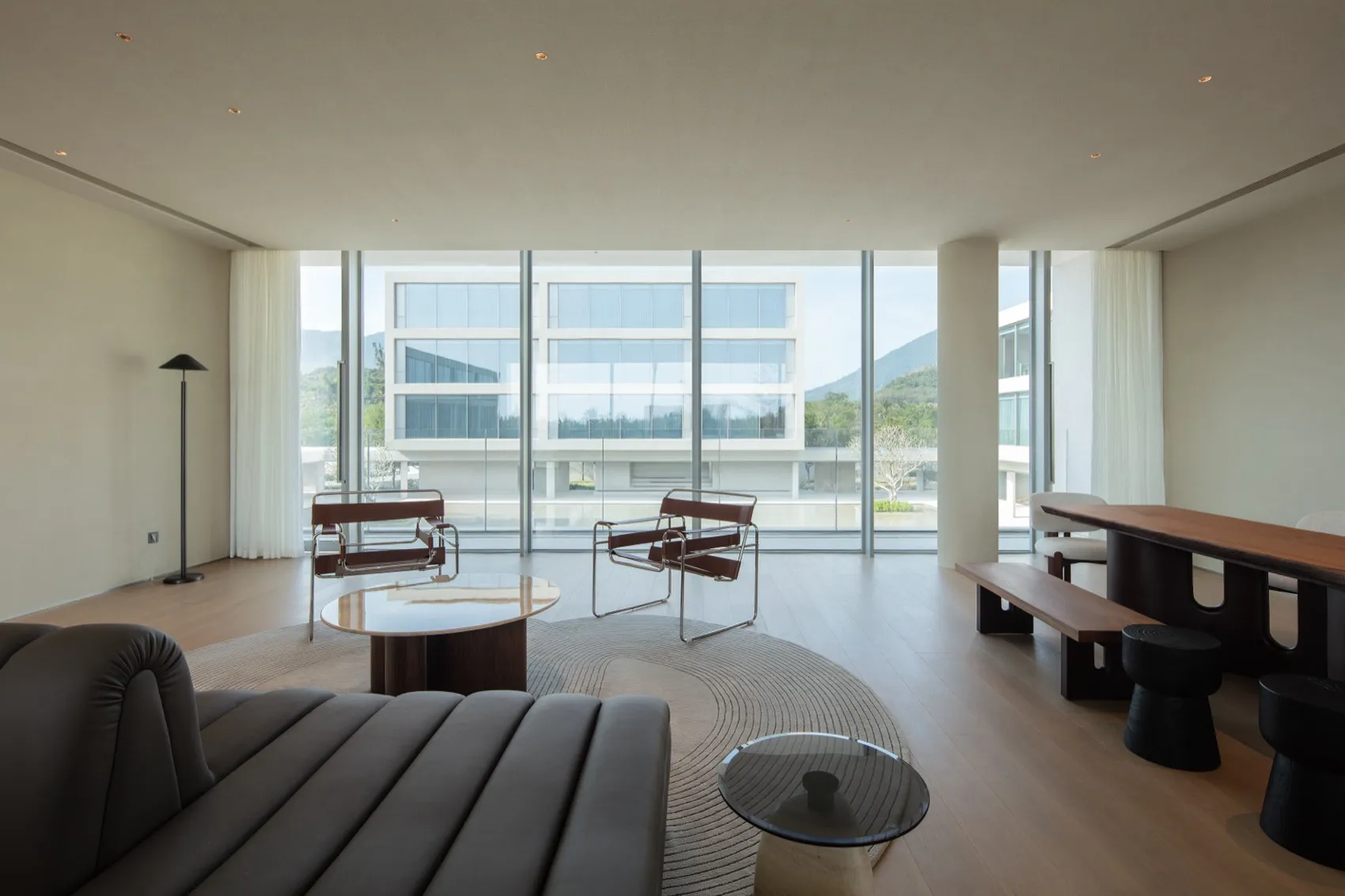 Modern minimalist living room featuring brown leather and chrome lounge chairs, walnut coffee tables, a curved rug, and large floor-to-ceiling windows overlooking mountains and contemporary buildings.