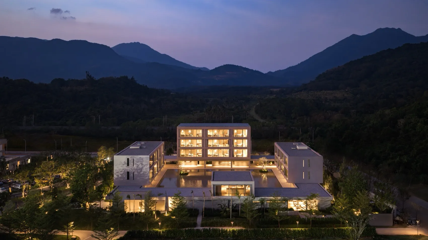 鳥瞰現代多棟度假酒店建築群，中央設有倒影池，在暮光中全亮照明，周圍環繞森林山脈。
