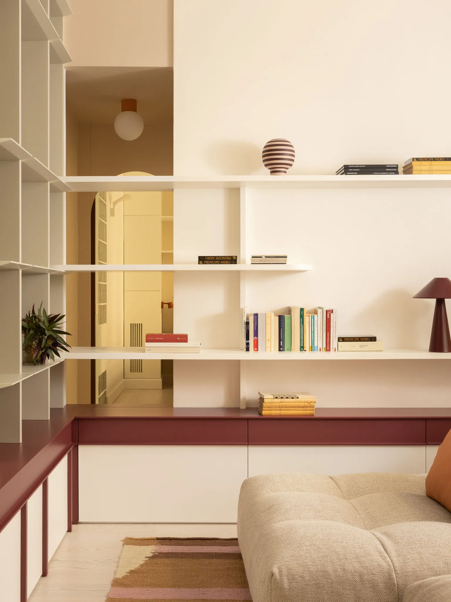 Modern minimalist living room featuring white floating shelves, burgundy lacquered cabinetry, a beige modular sofa, and warm lighting with a striped ceramic vase and colorful books.