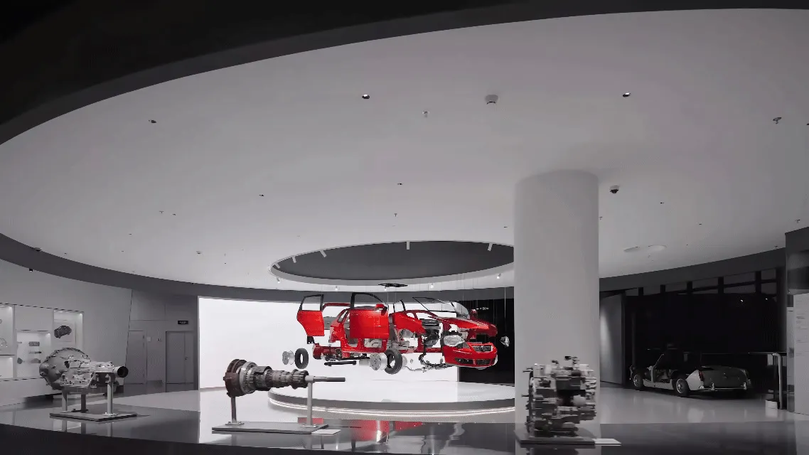 Modern exhibition hall with a red car chassis and body suspended in the center, surrounded by automotive components on stands under a curved white ceiling.