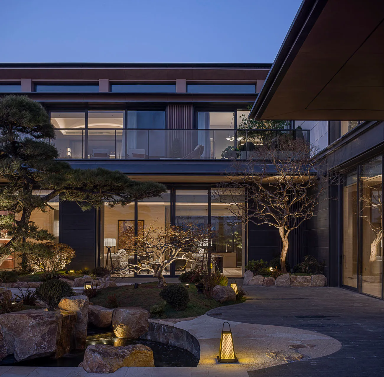 Modern Japanese-style courtyard at dusk featuring a stone-lined pond, pine tree, bare trees, stone path, and glowing lanterns against a contemporary house with large glass windows.
