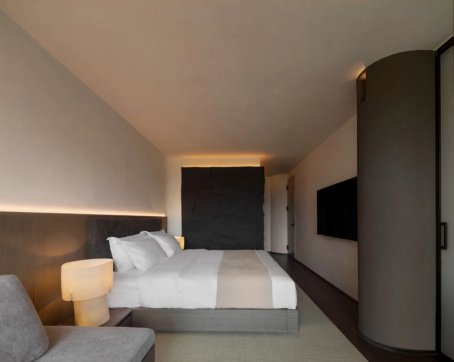 Modern minimalist bedroom interior with a large dark stone feature wall behind the bed, warm LED cove lighting, neutral bedding, and a curved dark wall panel on the right.