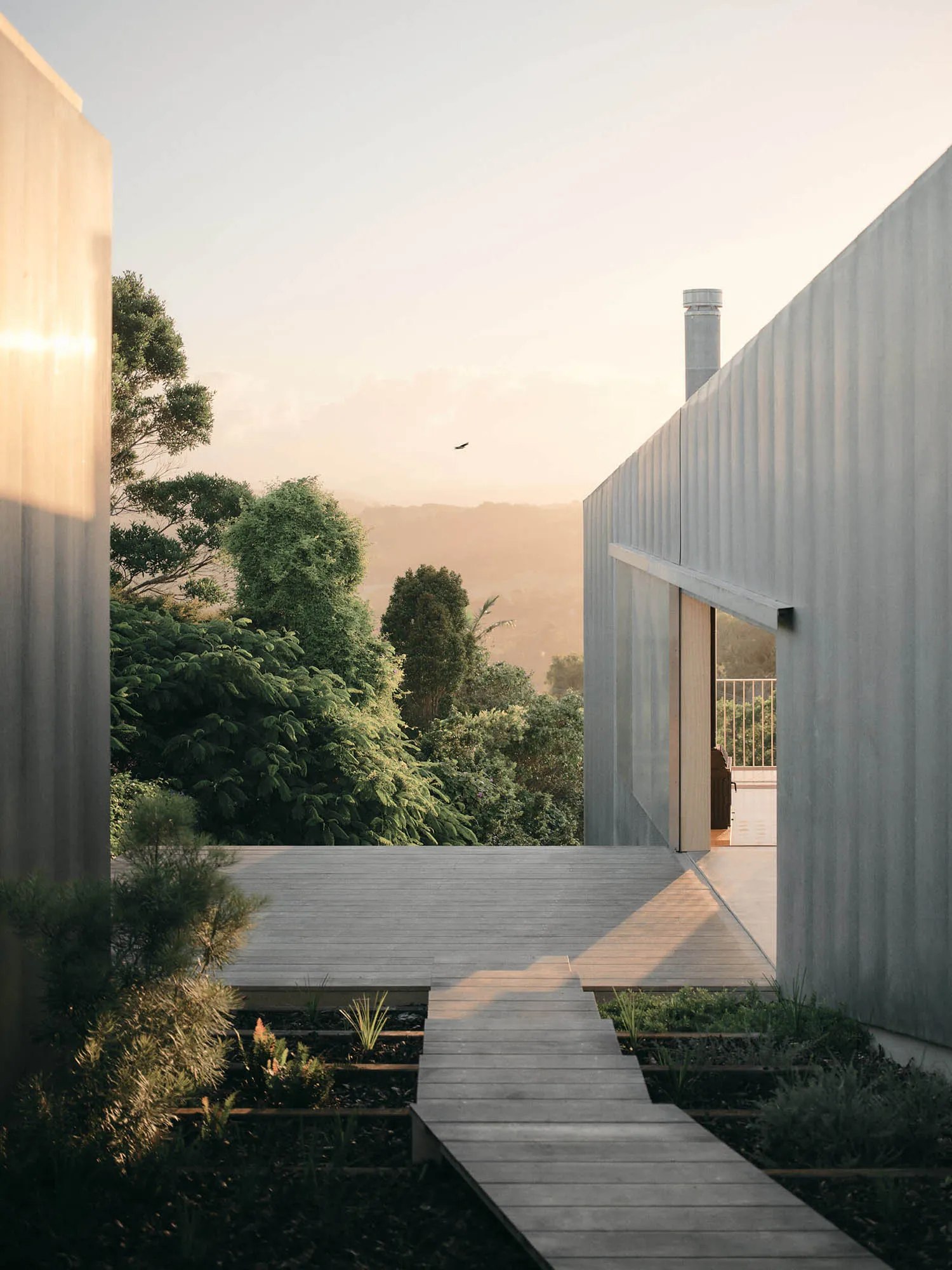 View from inside a modern concrete house looking out onto a wooden deck terrace at golden hour with forested hills in the background.