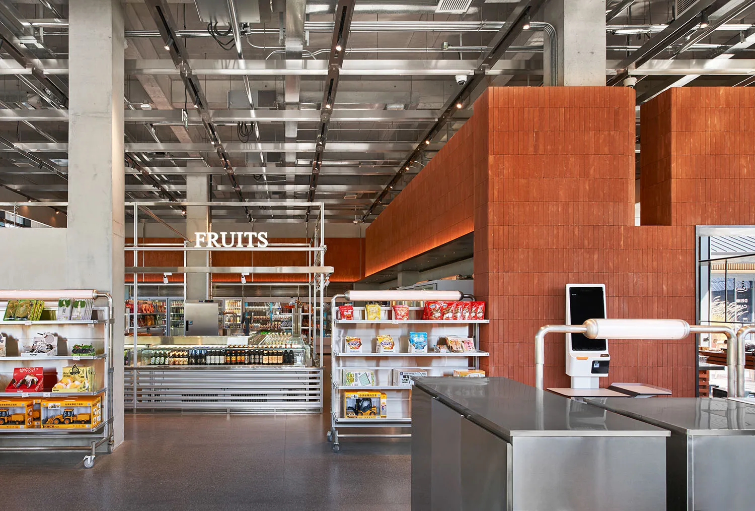 Modern supermarket interior showing stainless steel checkout counters, orange-brown corten steel wall panels, metal shelving units, and an exposed industrial ceiling with visible ductwork and lighting.