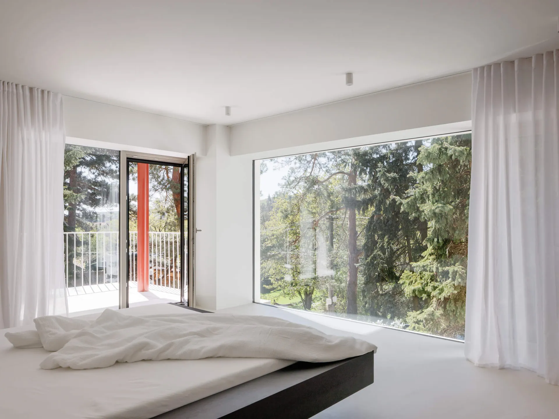 Minimalist bedroom with low platform bed dressed in white linen, sheer white curtains, and large windows overlooking a lush green forest.