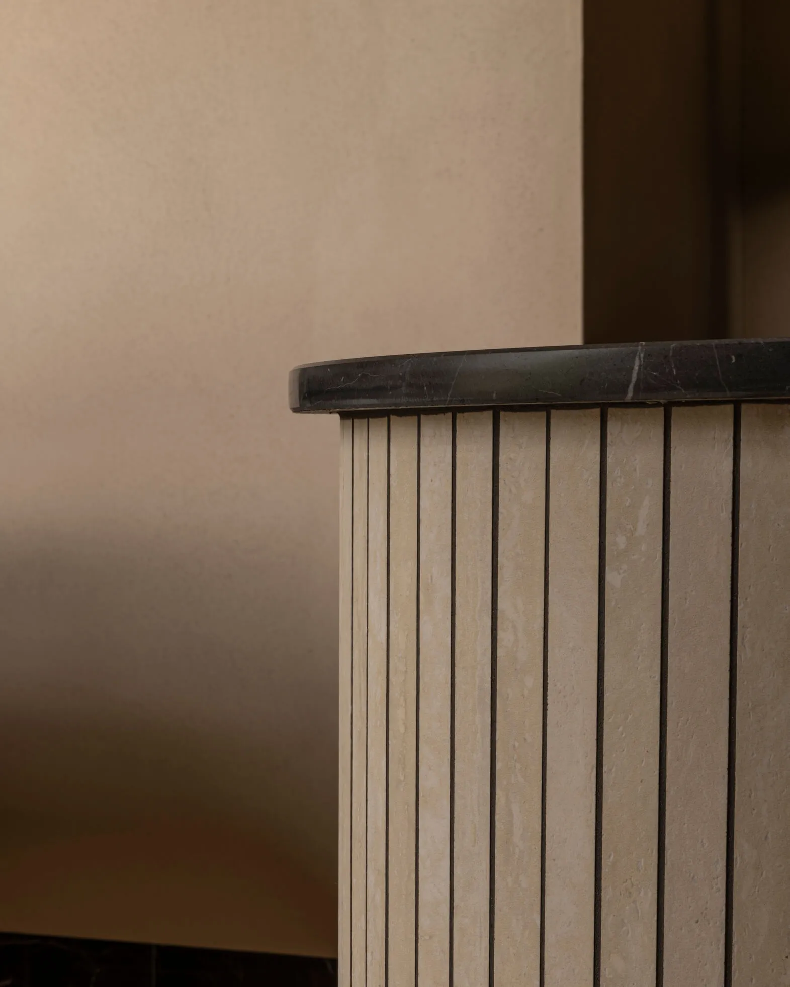 Close-up of a fluted travertine cabinet featuring vertical grooves and a dark marble countertop against a neutral textured wall.