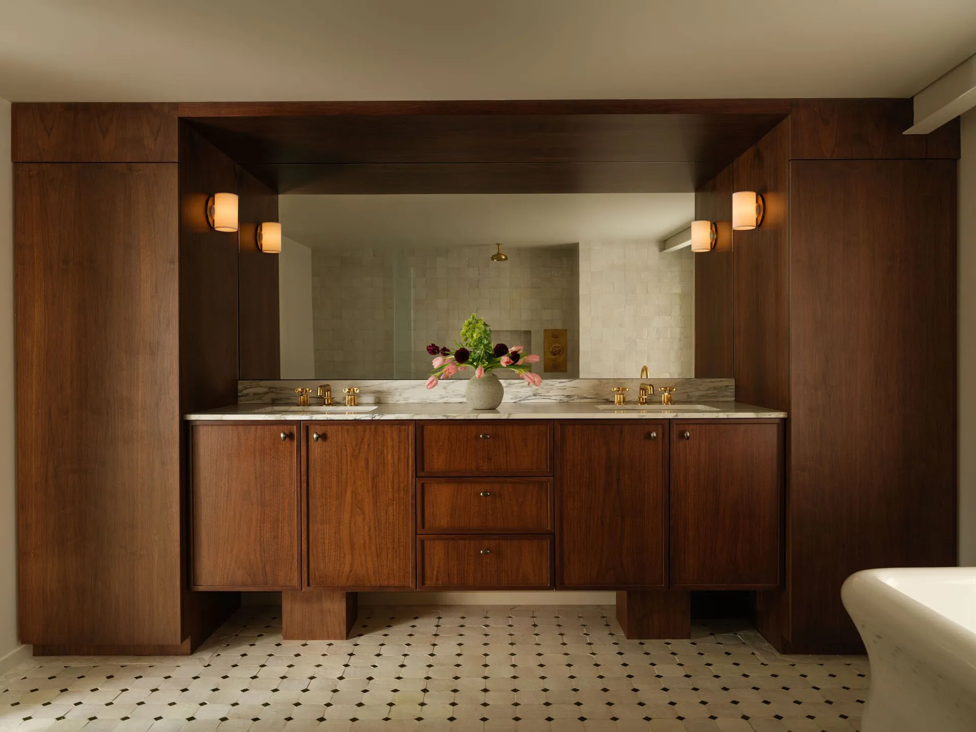 Luxurious bathroom with a double walnut vanity, white marble countertop and floor tiles, large mirror, and brass wall sconces.