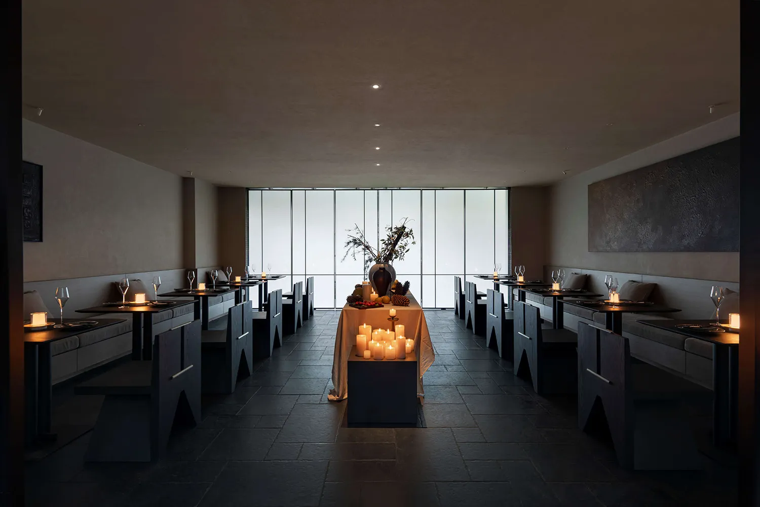 Interior view of a minimalist fine dining restaurant featuring dark stone floors and walls, built-in banquette seating, angular dark wood tables and chairs, and numerous lit candles creating warm ambient lighting, with large floor-to-ceiling windows at the far end.