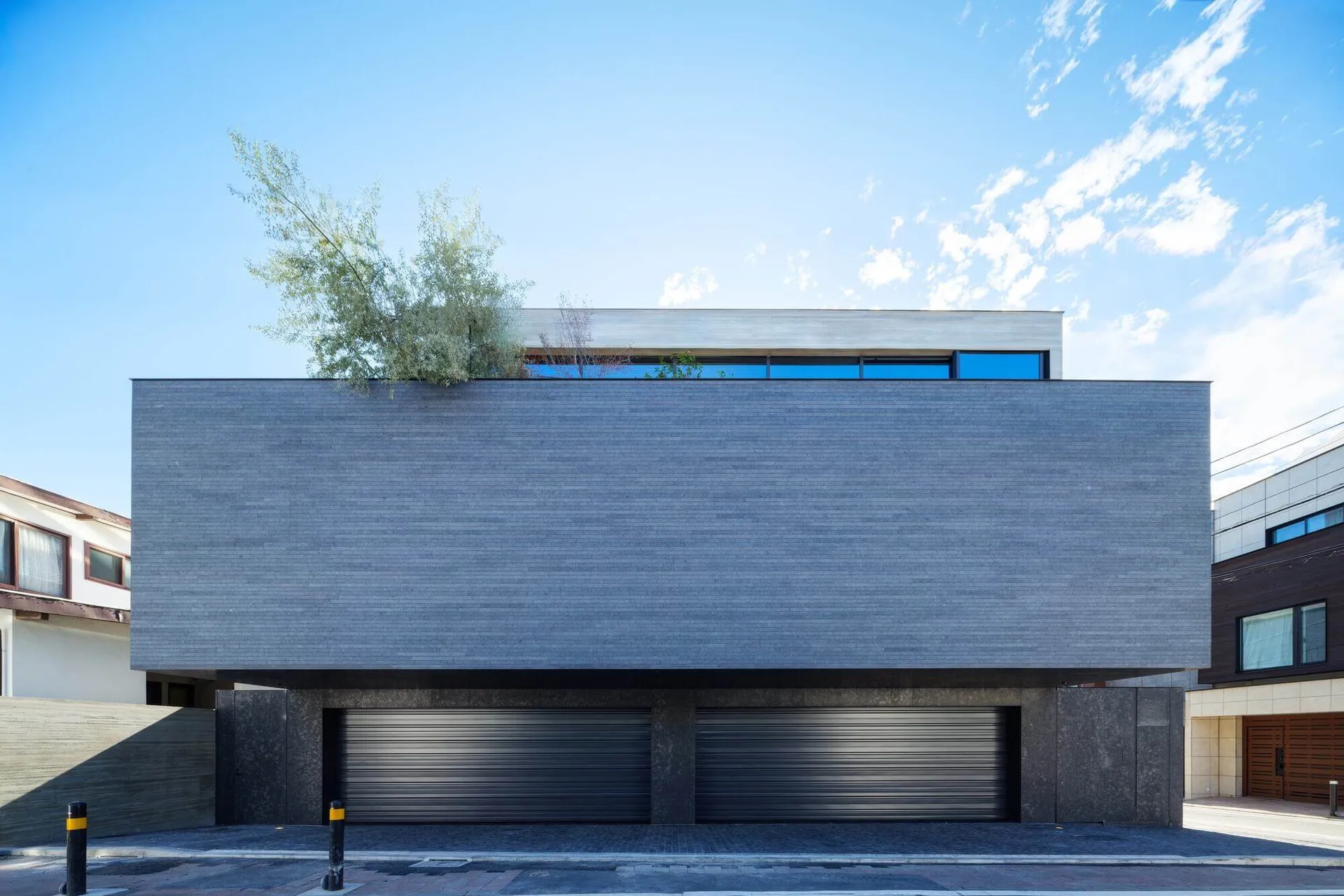 Modern brutalist residential building with textured grey concrete facade, two dark roller garage doors, and a rooftop garden with trees under a clear blue sky.