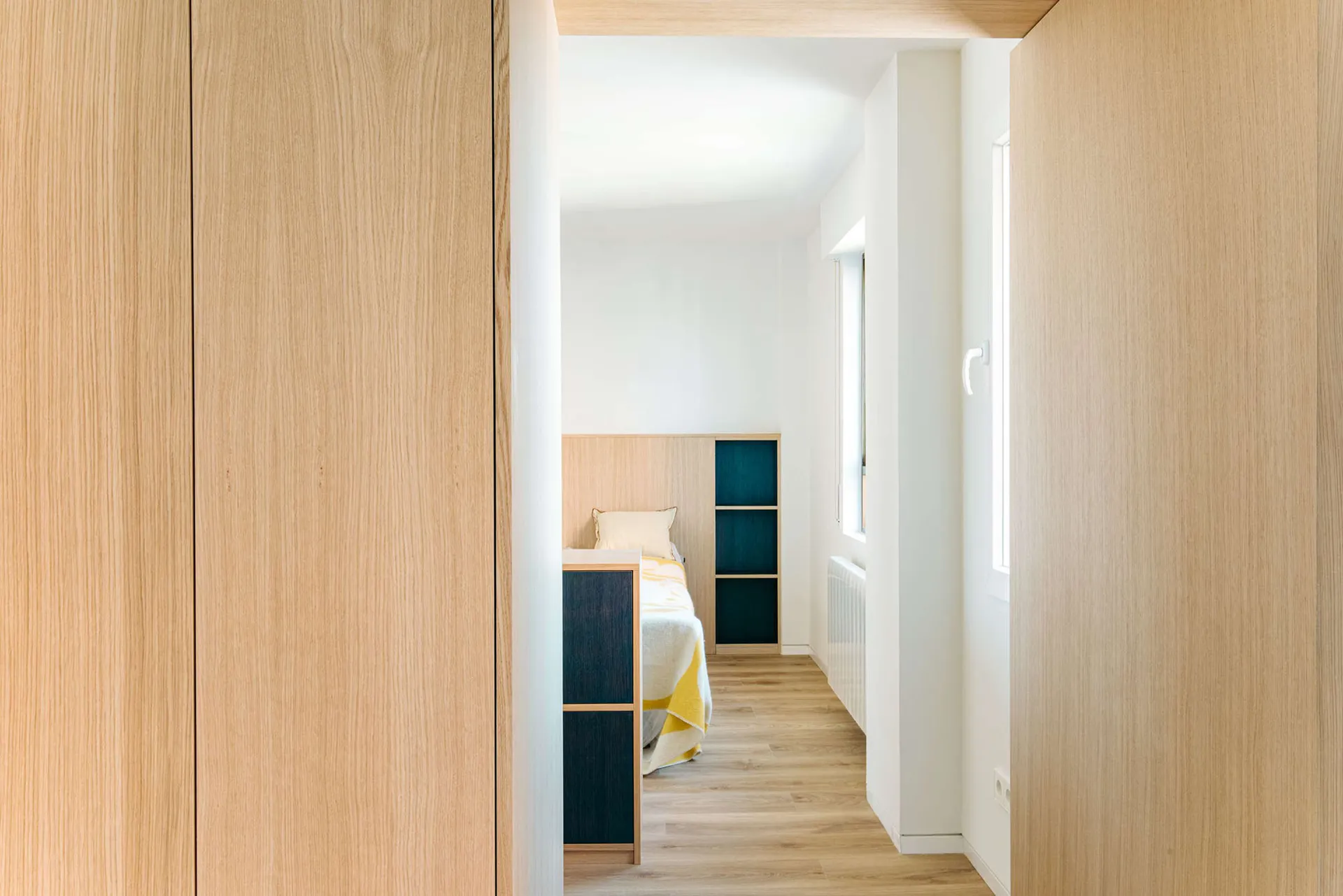View from a doorway into a narrow modern bedroom with light oak wood paneling, built-in teal storage units, and a bed with yellow and white bedding.