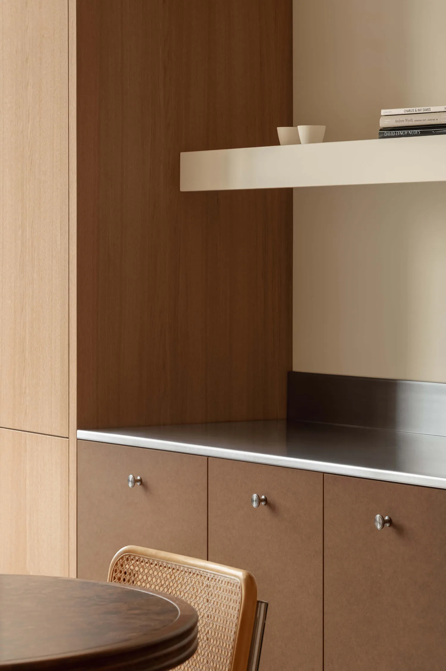 Close-up of a modern dining area featuring warm walnut lower cabinets with round silver knobs, a stainless steel countertop, light oak upper cabinetry, and a cane-backed dining chair in the foreground.