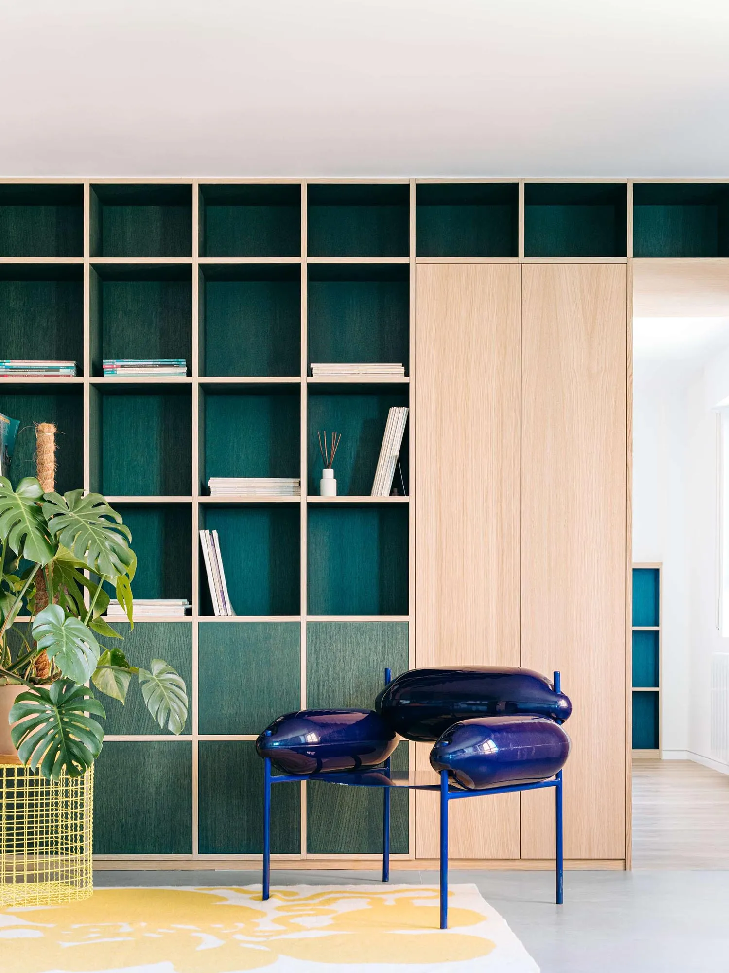 Modern living room with teal velvet built-in shelving, light wood cabinetry, a glossy blue sculptural chair, a large monstera plant, and a yellow floral rug.