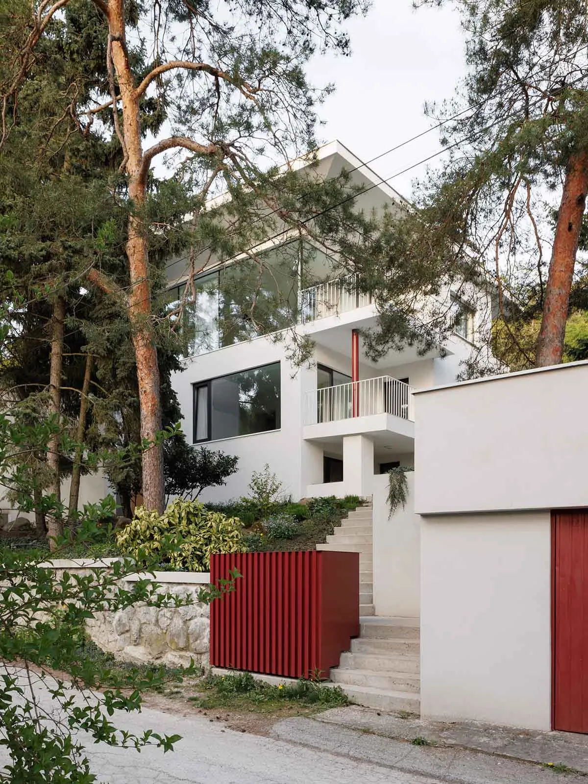 Modern white house with large glass windows, white balconies, concrete stairs, and vibrant red metal accents on a wooded hillside.