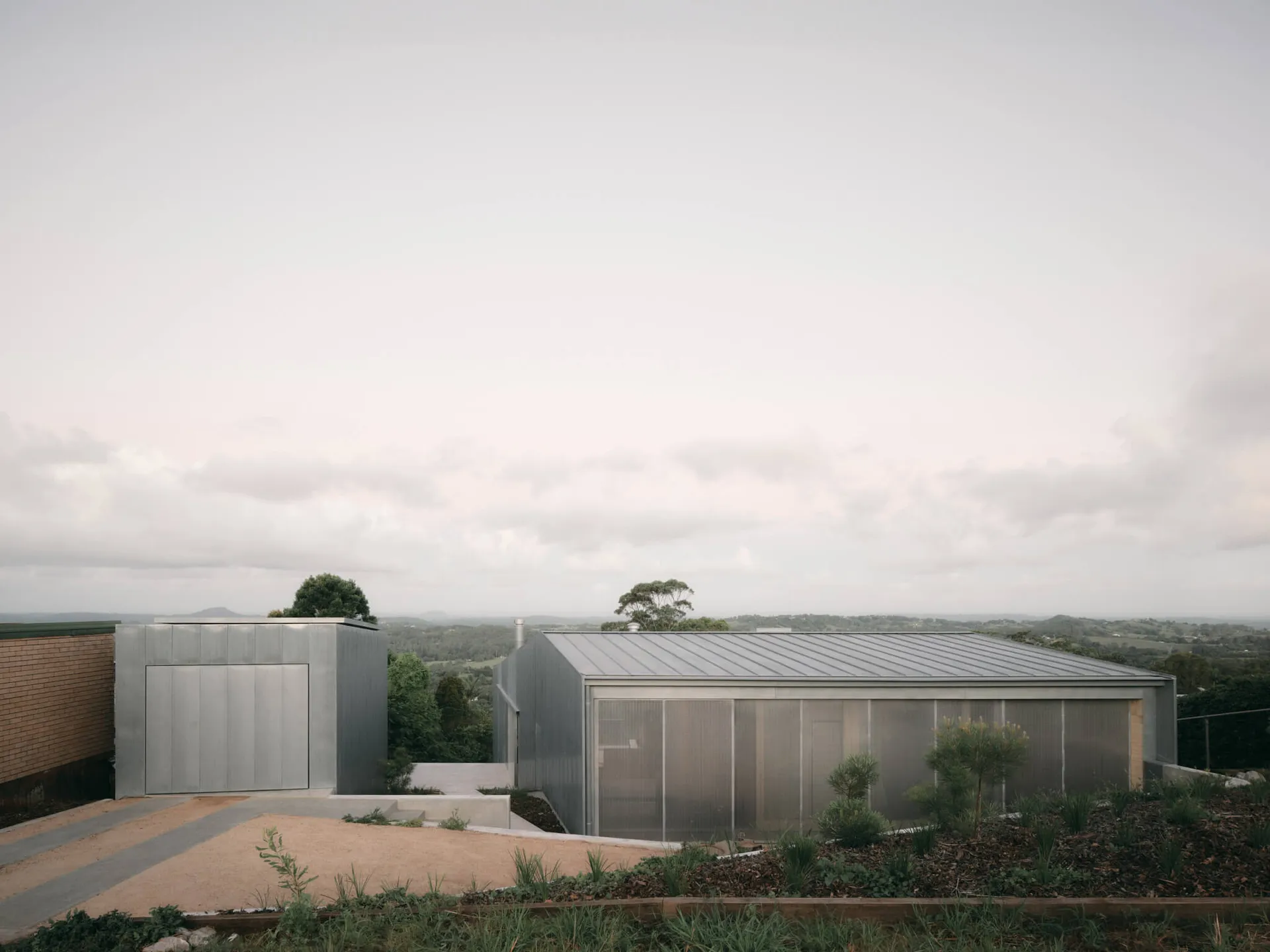 Modern single-story house with corrugated metal cladding and large translucent windows, situated on a hilltop overlooking a rural valley landscape under an overcast sky.