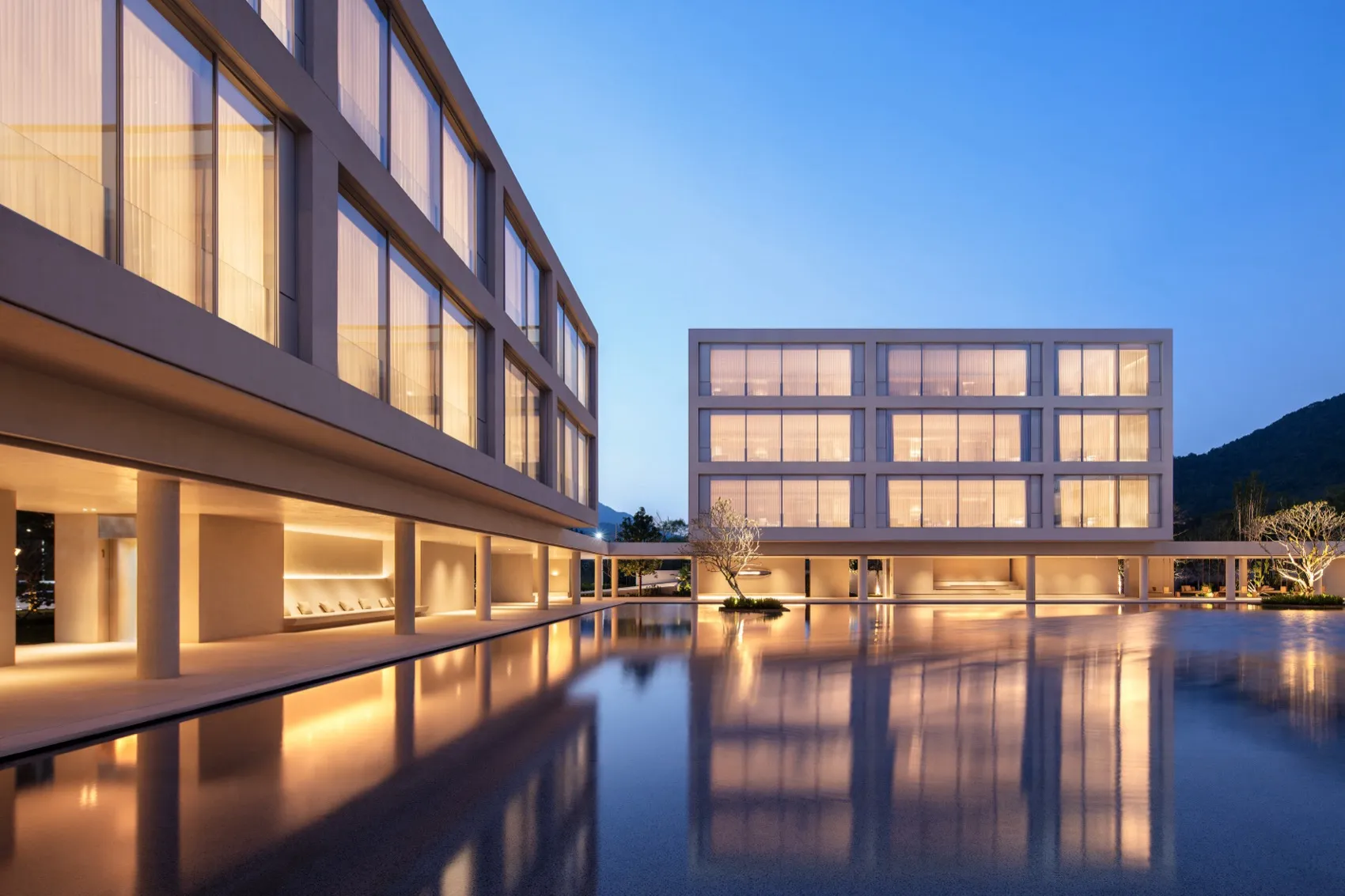 Modern minimalist building complex with large glass windows and concrete structure surrounding a large rectangular reflection pool at twilight, with mountains in the background.
