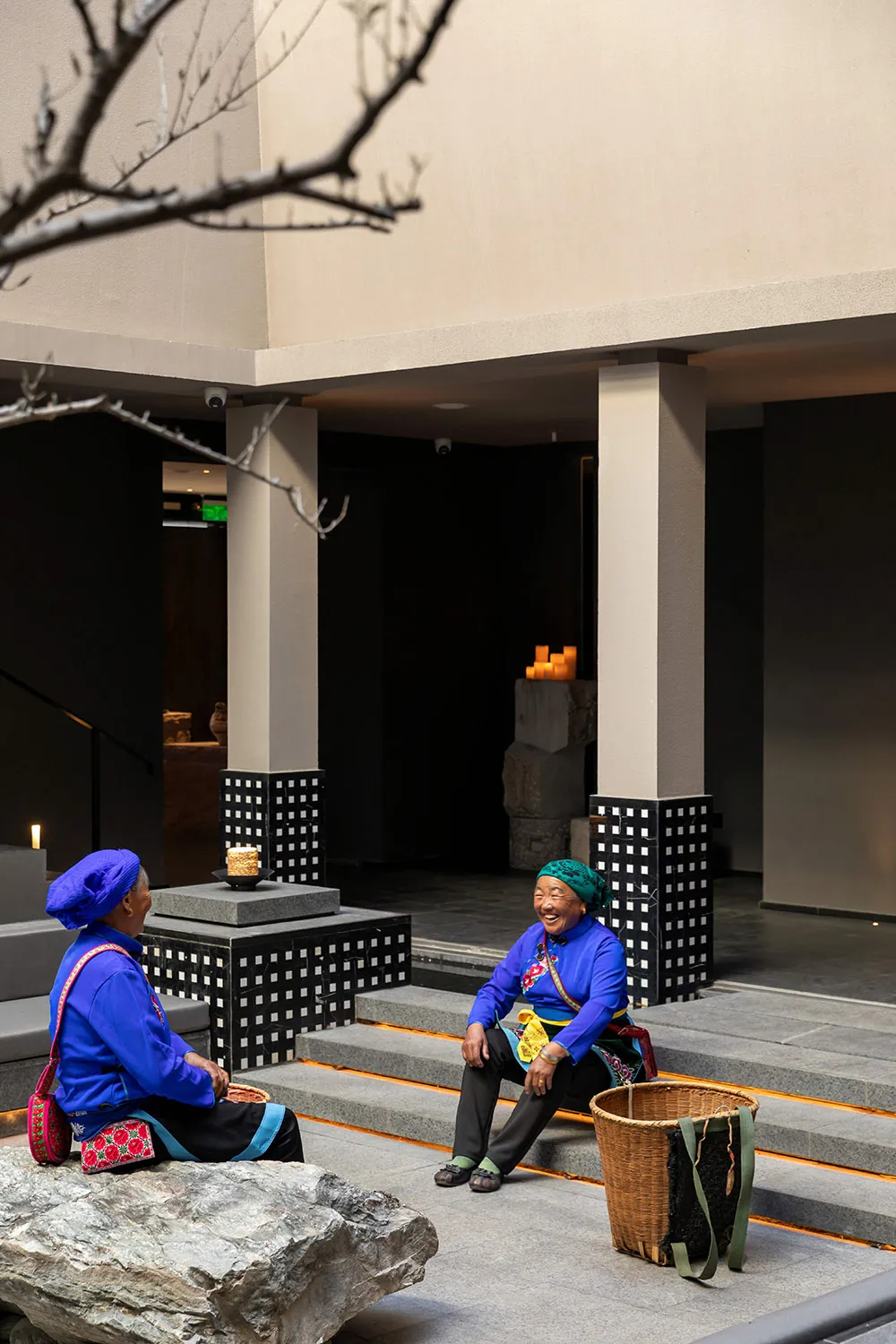 Two elderly Miao women wearing vibrant blue traditional costumes and headwraps sit on stone steps in a modern concrete and stone courtyard at night, illuminated by warm lighting.