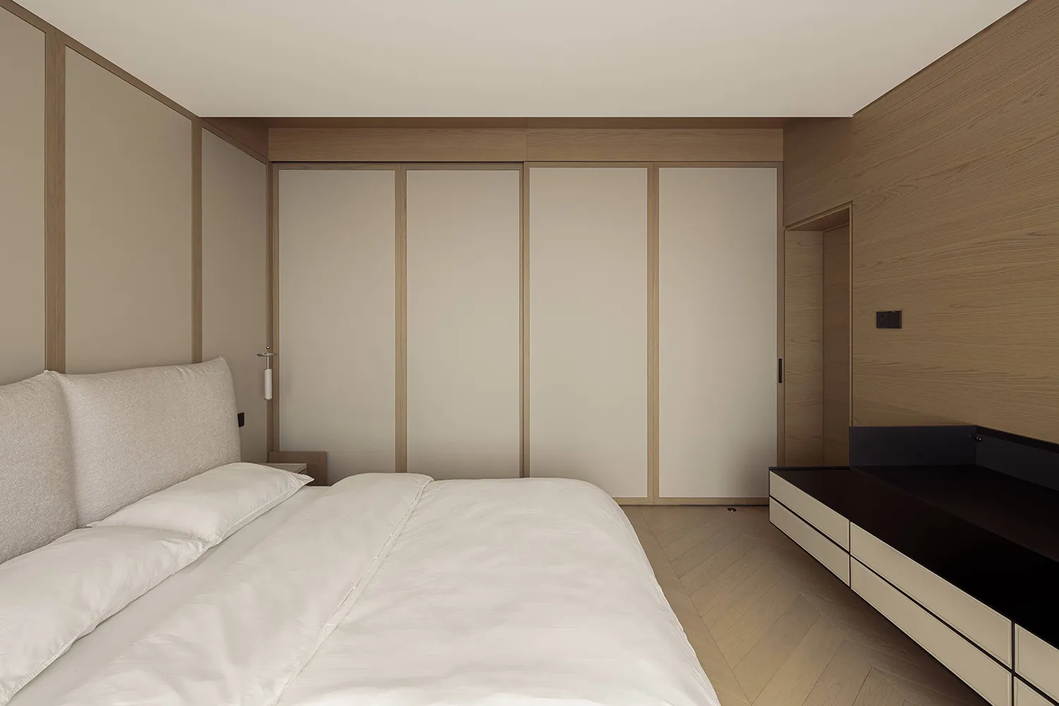 Modern minimalist bedroom with white bedding, upholstered headboard, oak veneer walls and wardrobe, and a black floating console on the right.