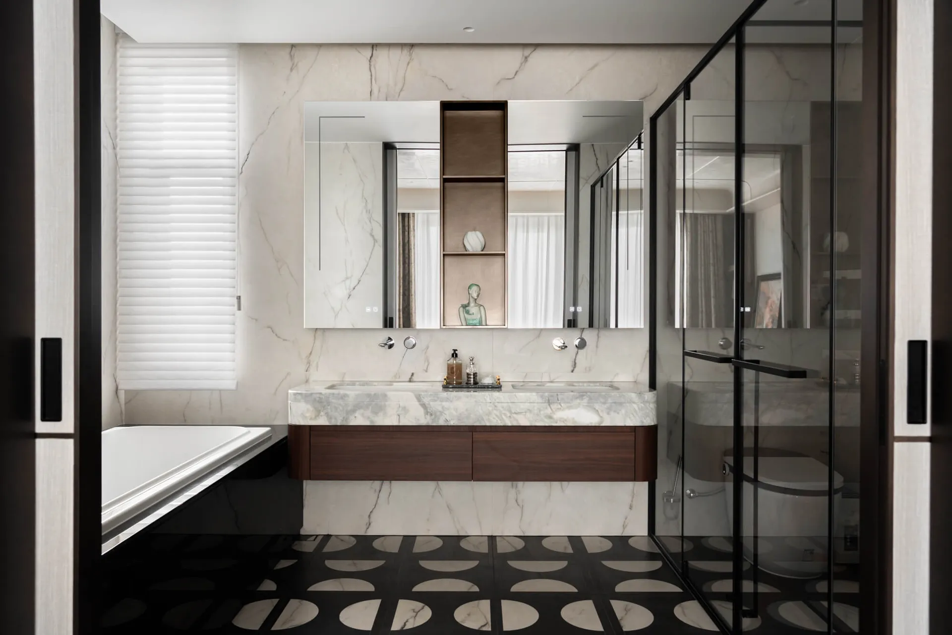 Modern luxury bathroom with white marble walls, dark wood floating vanity with integrated marble countertop and double sinks, black and white geometric patterned floor tiles, freestanding bathtub, and glass-enclosed shower.