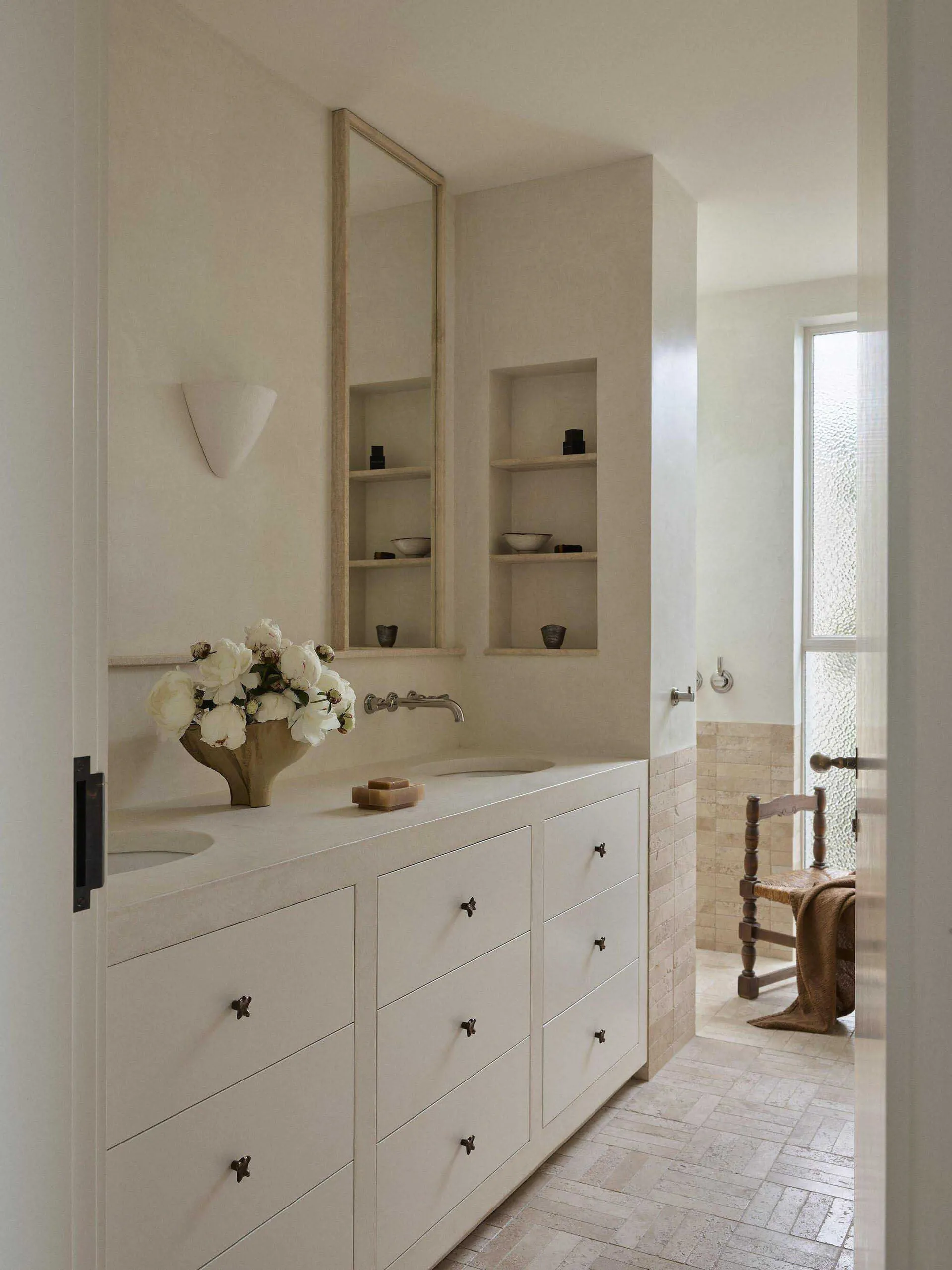 Modern minimalist bathroom with white limestone vanity, integrated sink, dark metal hardware, recessed wall niches, and a wooden chair visible in the adjacent space.
