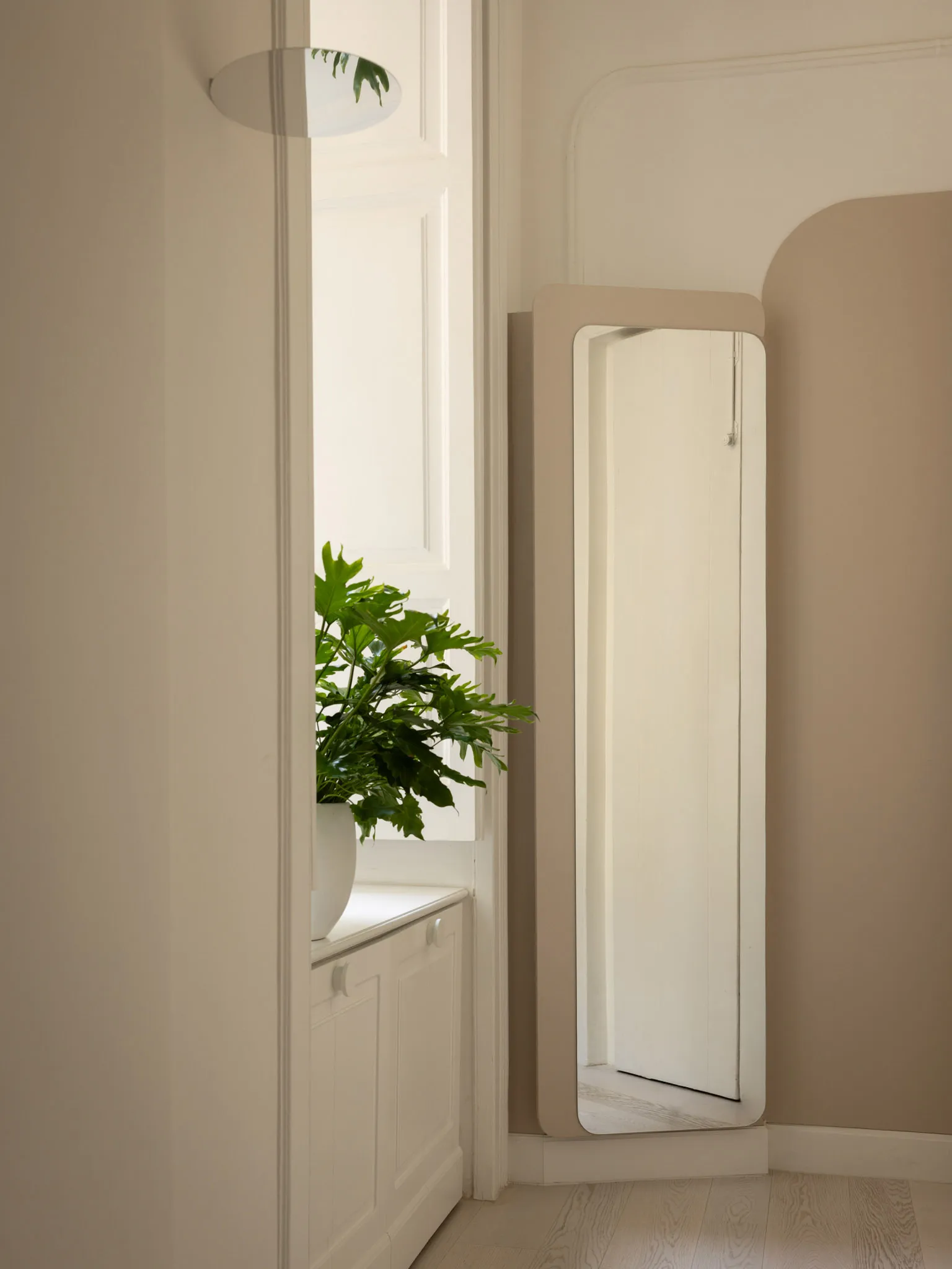 Minimalist entryway corner featuring a tall full-length mirror with a matte beige frame, a white arched wall detail, and a large potted monstera plant on a white cabinet by the window.