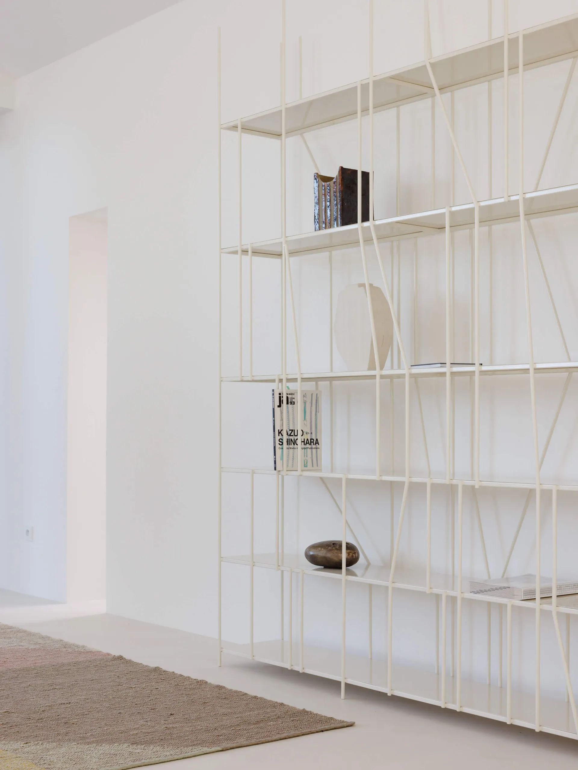 Modern white metal grid bookshelf against a white wall, displaying books, ceramic vases, and sculptural objects, with a textured area rug in the foreground.