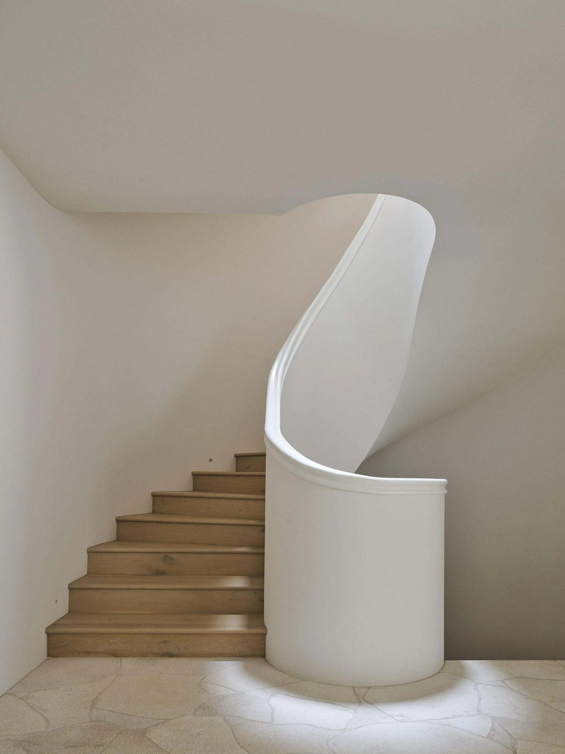 Modern minimalist interior featuring a curved white structural wall beside a wooden staircase with light oak treads and stone tile floor.