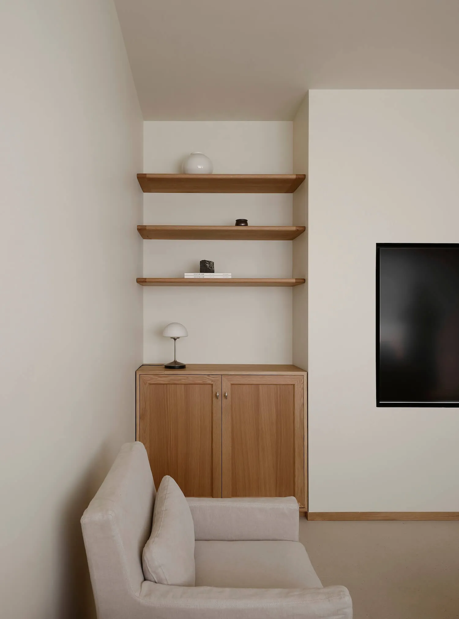Minimalist living room corner with light oak cabinet, three floating oak shelves, white walls, and a cream fabric sofa in the foreground.