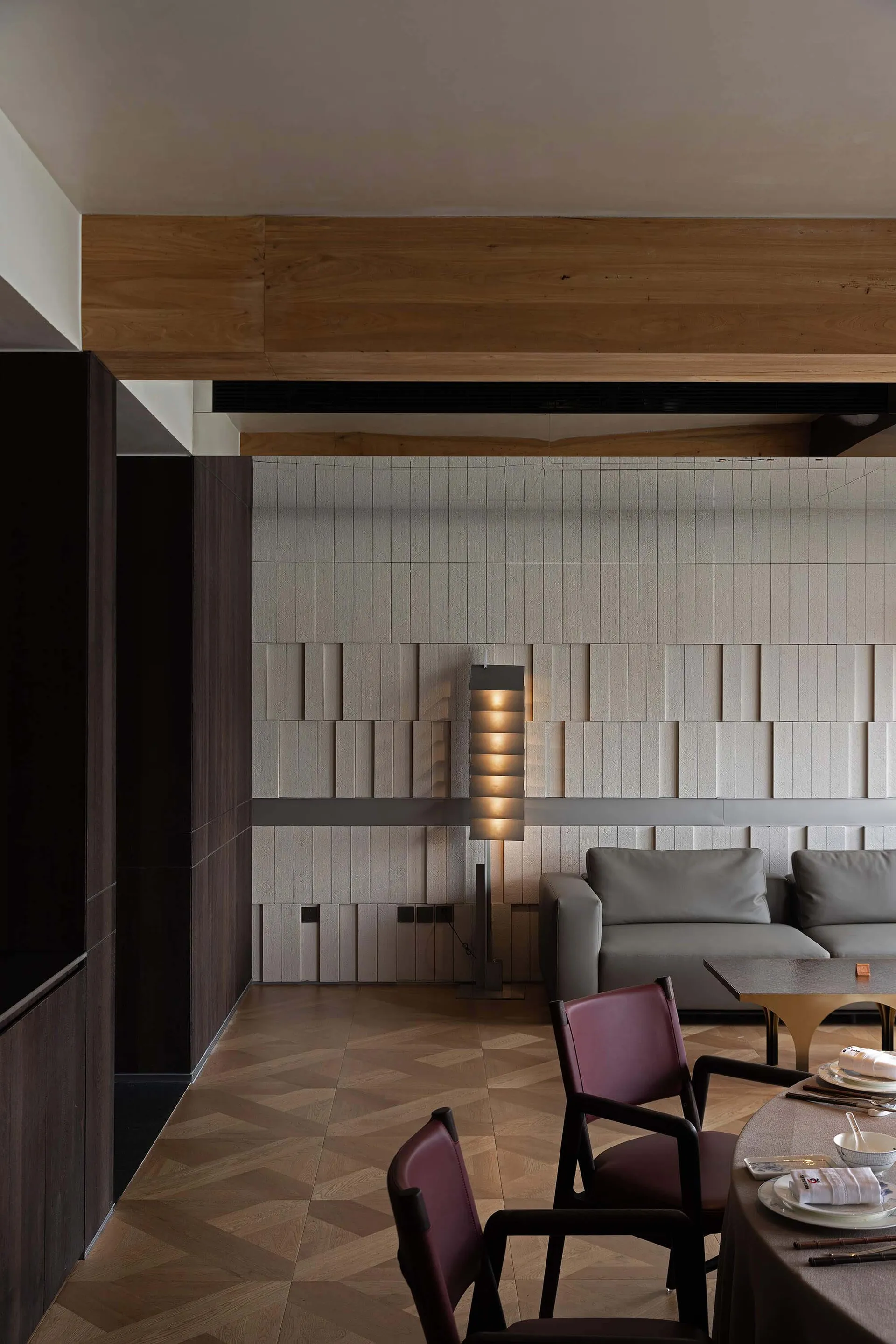 Modern dining room featuring burgundy leather chairs, a round dining table set with tableware, a grey sofa, herringbone wood flooring, and a feature wall with beige textured tiles and a wooden beam overhead.