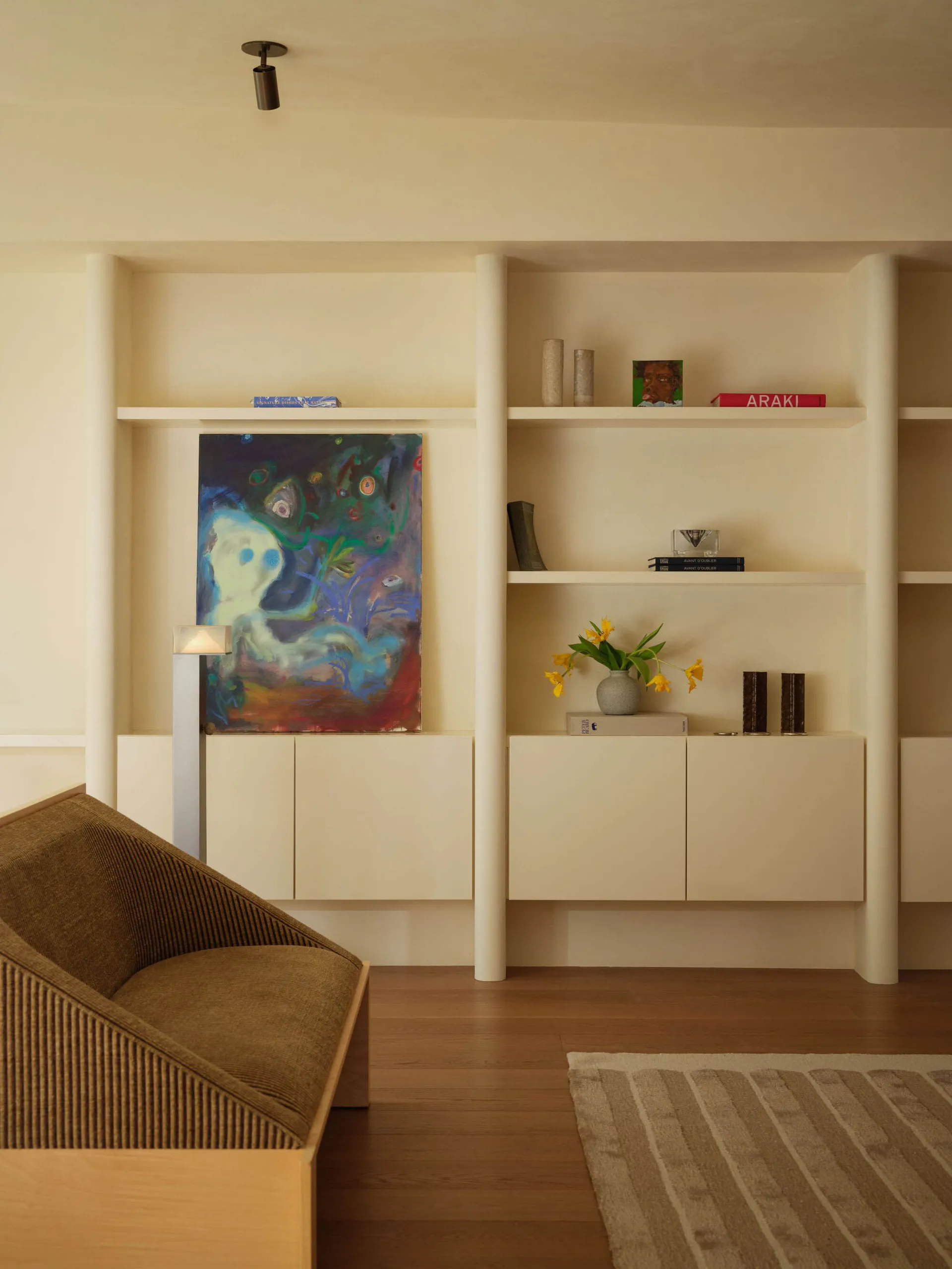 Modern minimalist living room with light oak flooring, built-in white shelving, a brown ribbed fabric armchair, colorful abstract painting, and decorative objects.