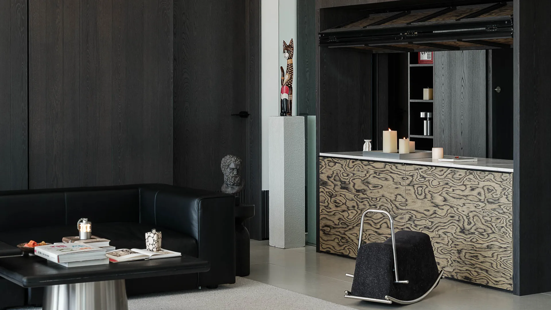 Modern dark living room featuring a black leather sofa, burl wood cabinet with white marble top, black rocking ottoman, concrete pedestal with cat sculpture, and dramatic dark wood paneling.