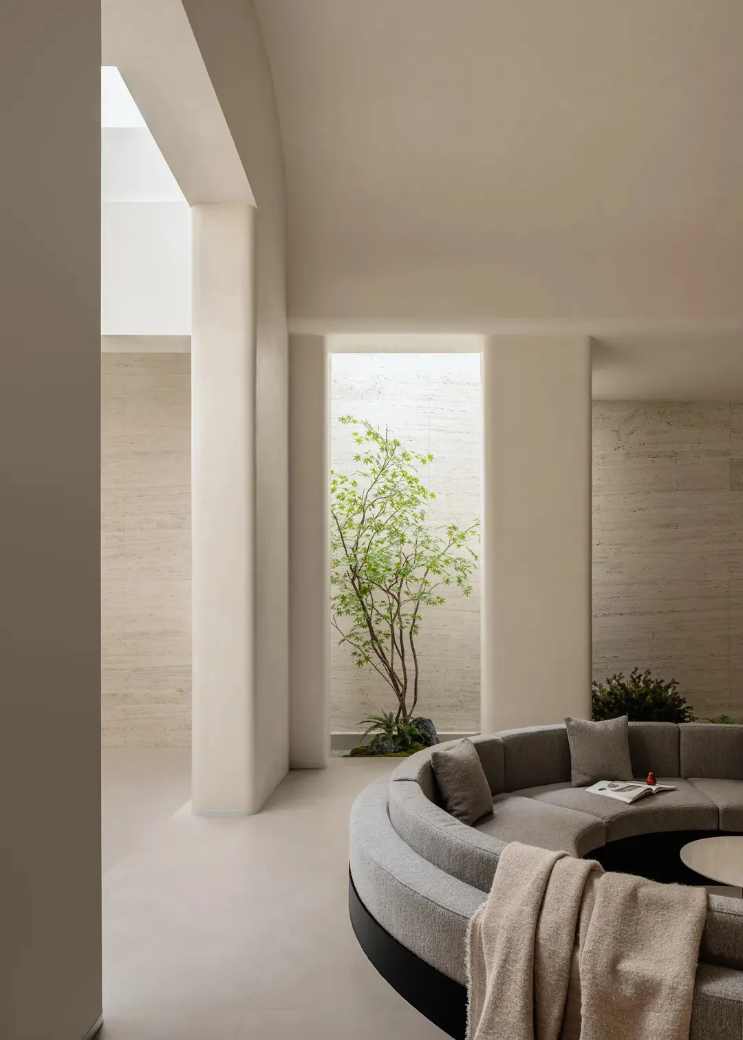 Modern minimalist living room featuring a large curved grey sectional sofa, travertine stone walls and floor, and an indoor courtyard with a Japanese maple tree.