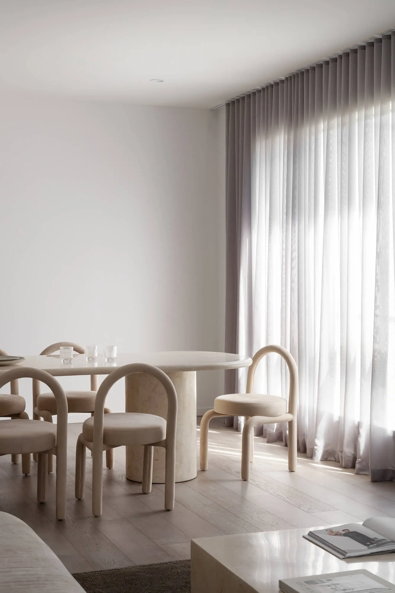 Minimalist dining room featuring a round travertine table, curved beige upholstered chairs, light wood flooring, and floor-to-ceiling sheer curtains with bright natural light.