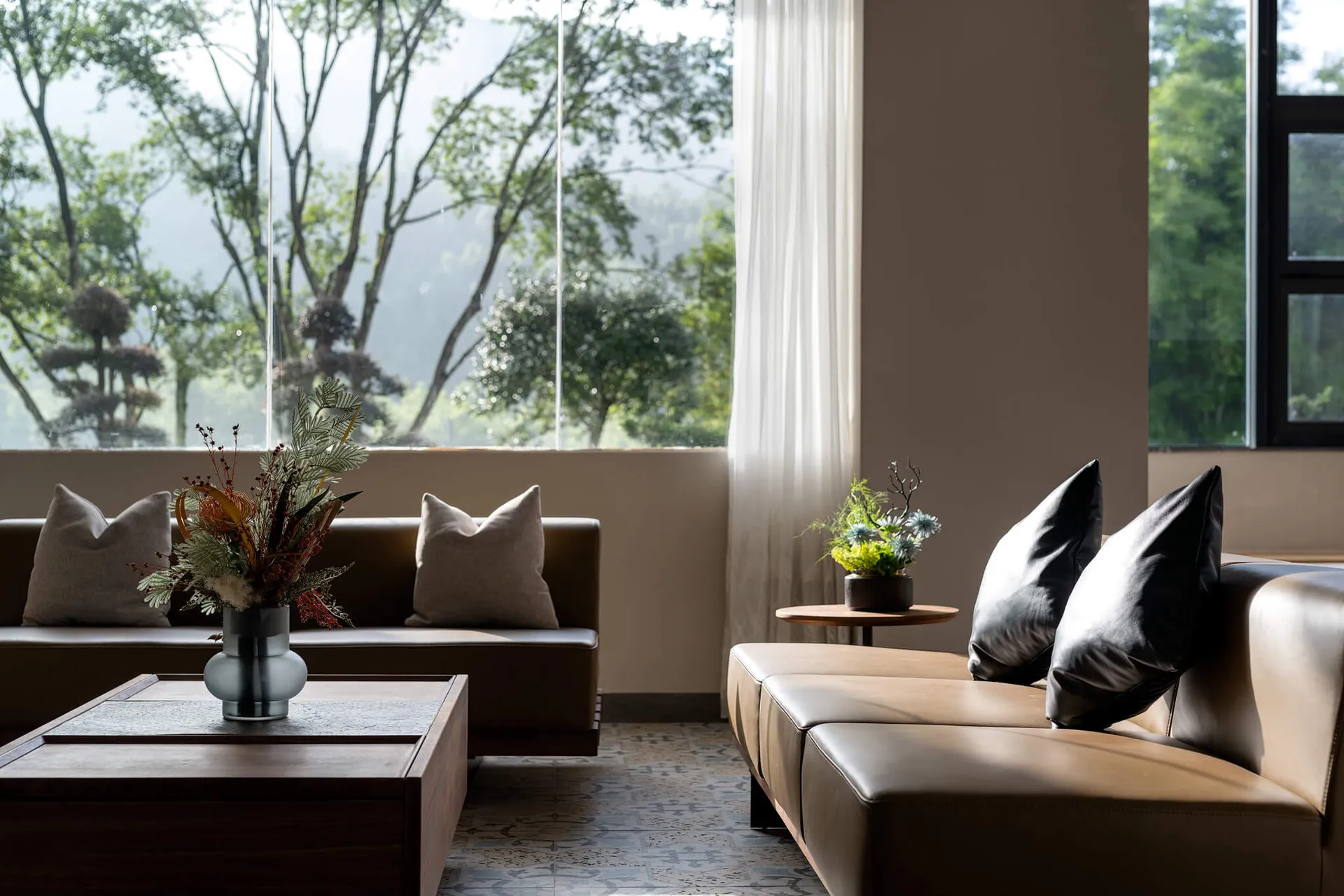 現代風客廳,棕色皮革沙發搭配黑色抱枕,胡桃木茶几上擺放金屬花瓶與乾燥花束,大面窗景可眺望鬱鬱蔥蔥的山林。