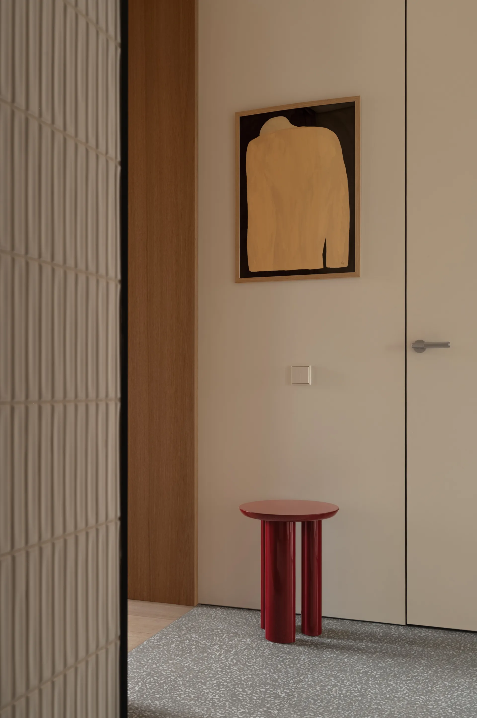 Modern minimalist entryway featuring a glossy red cylindrical side table on a grey terrazzo floor, with a large abstract painting in warm tones hanging on a beige wall beside a wooden door and vertical ribbed wall panel.