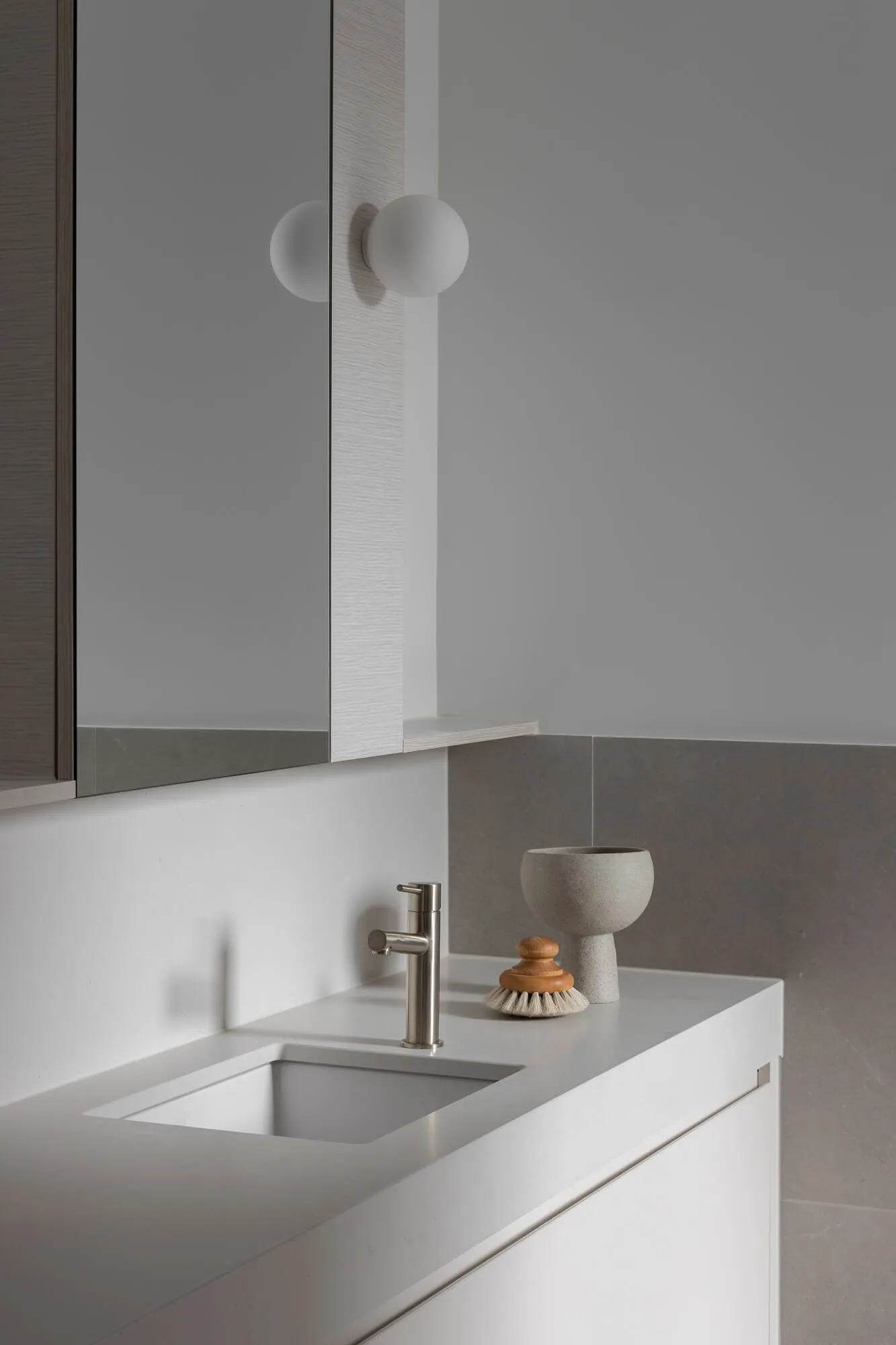Modern minimalist bathroom vanity featuring a white integrated sink, brushed nickel faucet, concrete bowl, wooden brush, and two spherical wall lamps against grey tiled walls.