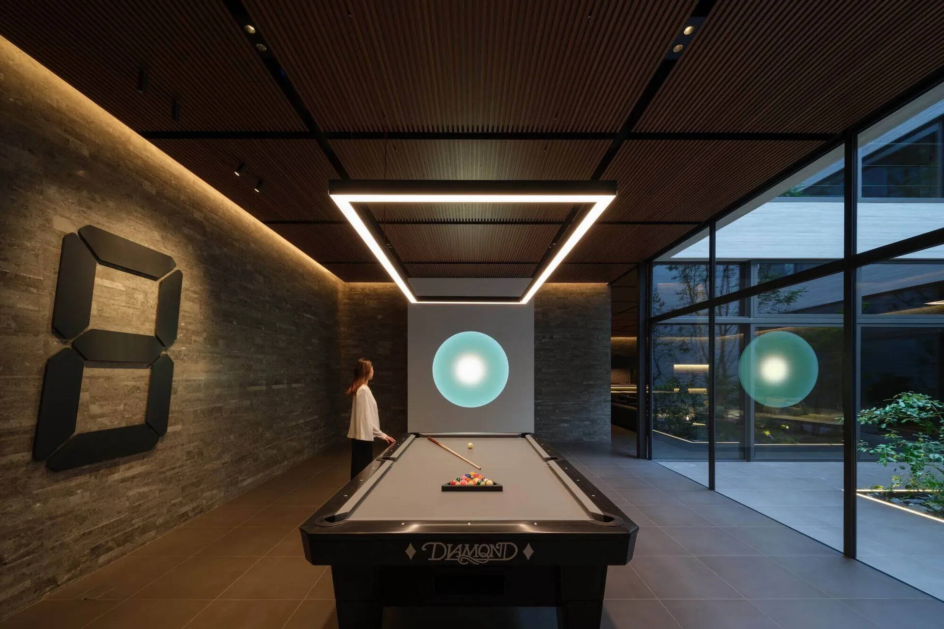 Modern minimalist billiard room with dark stone accent wall, black pool table, large square LED light fixture, and glowing cyan spherical pendant lights.