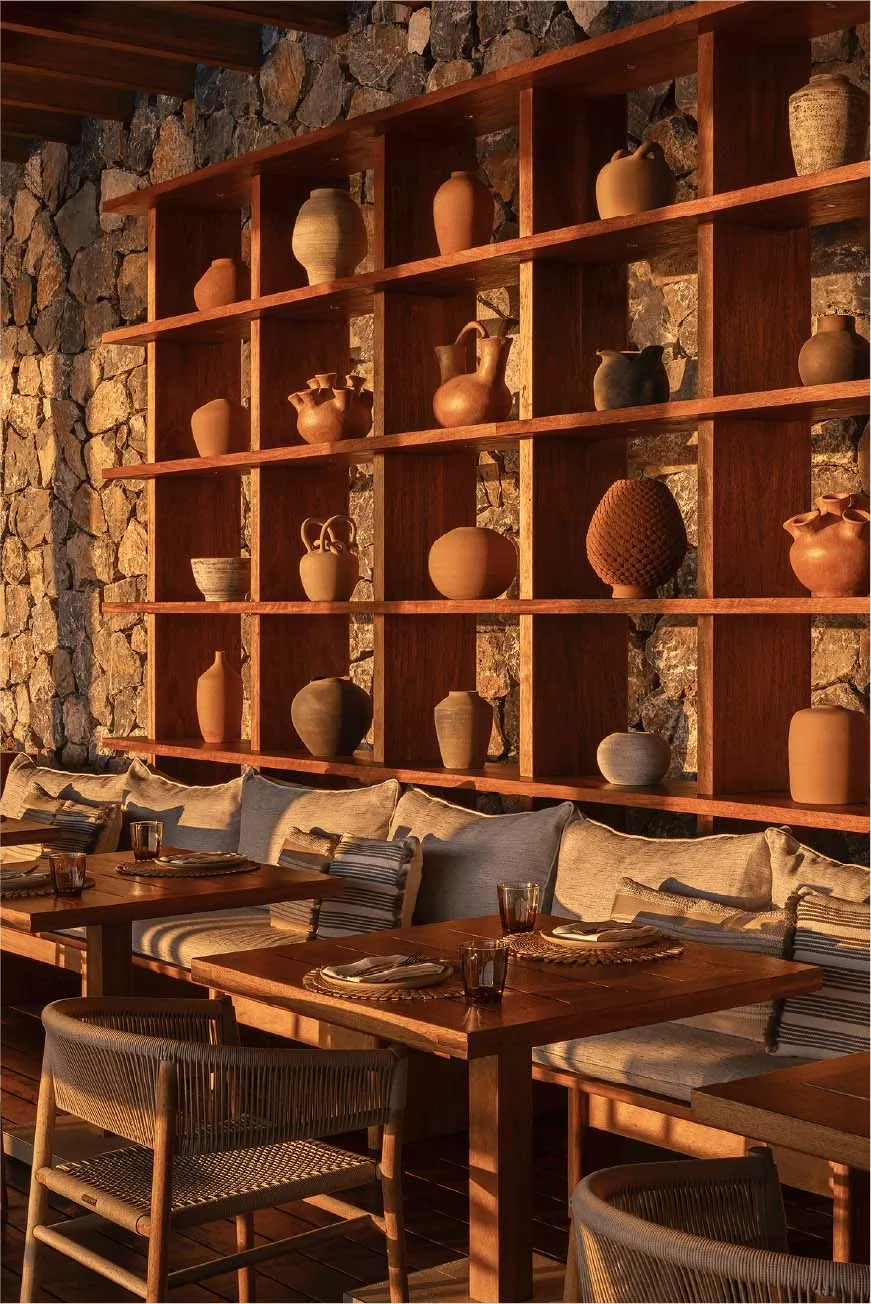 Cozy rustic dining area featuring a built-in wooden shelving unit filled with terracotta pottery, set against a natural stone wall with cushioned banquette seating and woven chairs.