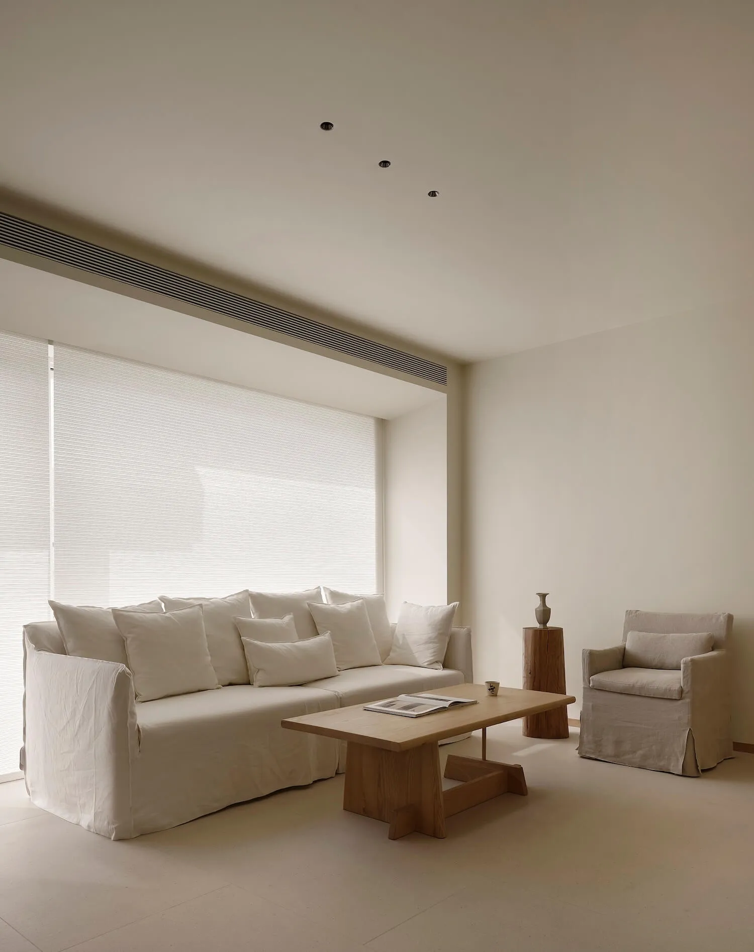 Minimalist living room featuring a large white linen sectional sofa, matching armchair, solid oak coffee table, and wooden side table against a backdrop of large windows with sheer blinds.