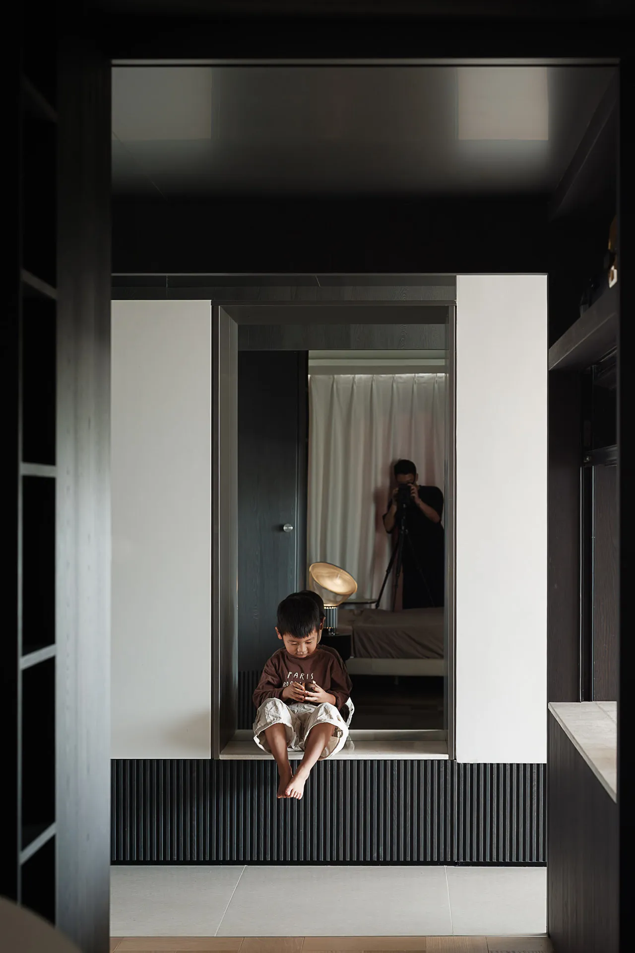 A young boy sitting barefoot on a modern built-in window seat with dark fluted wood paneling below, white walls, and a photographer visible in the reflection of a mirror.