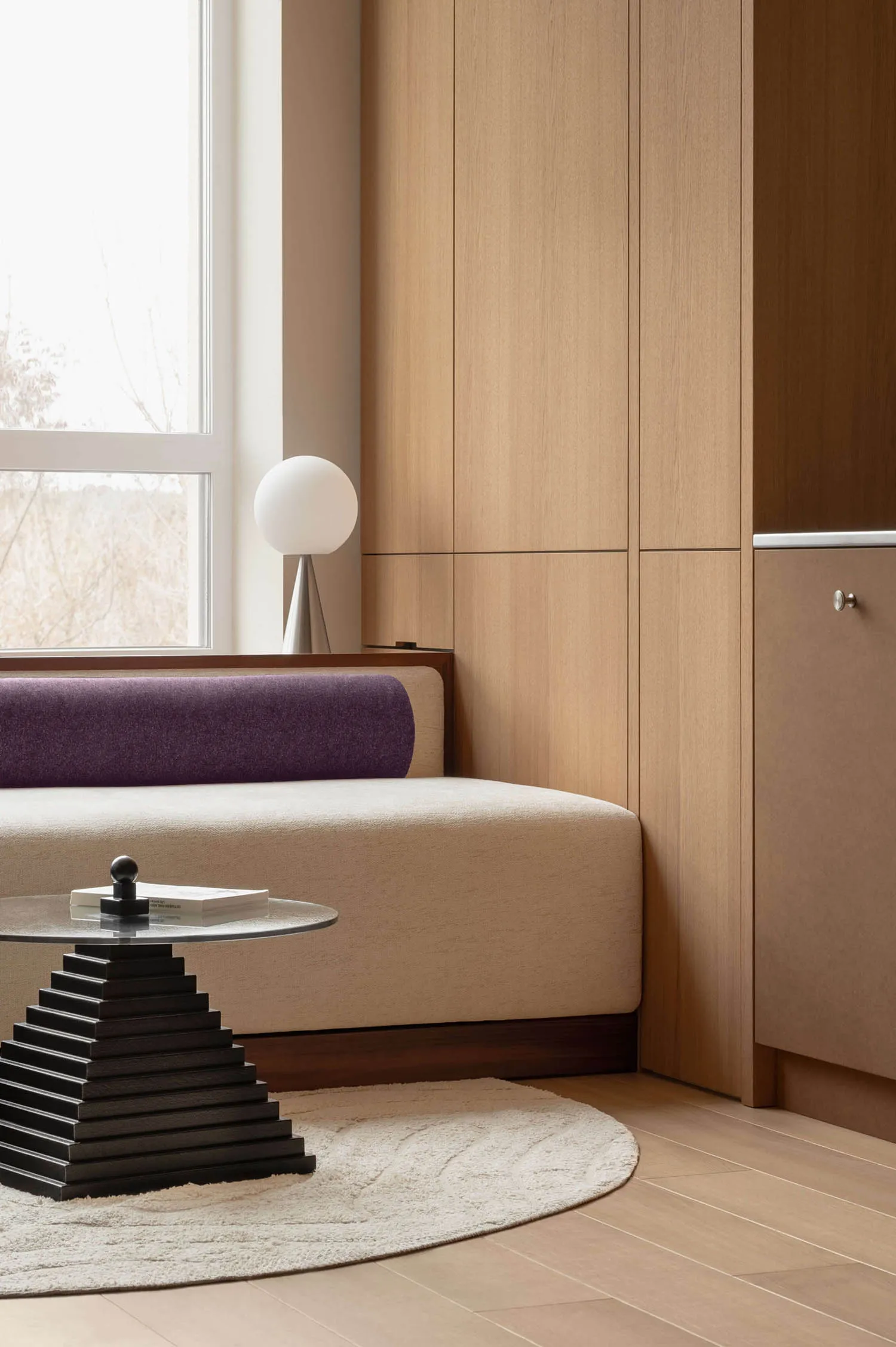 Modern living room corner featuring a purple upholstered window seat, light oak built-in storage, spherical table lamp, and a black geometric coffee table on a cream rug.