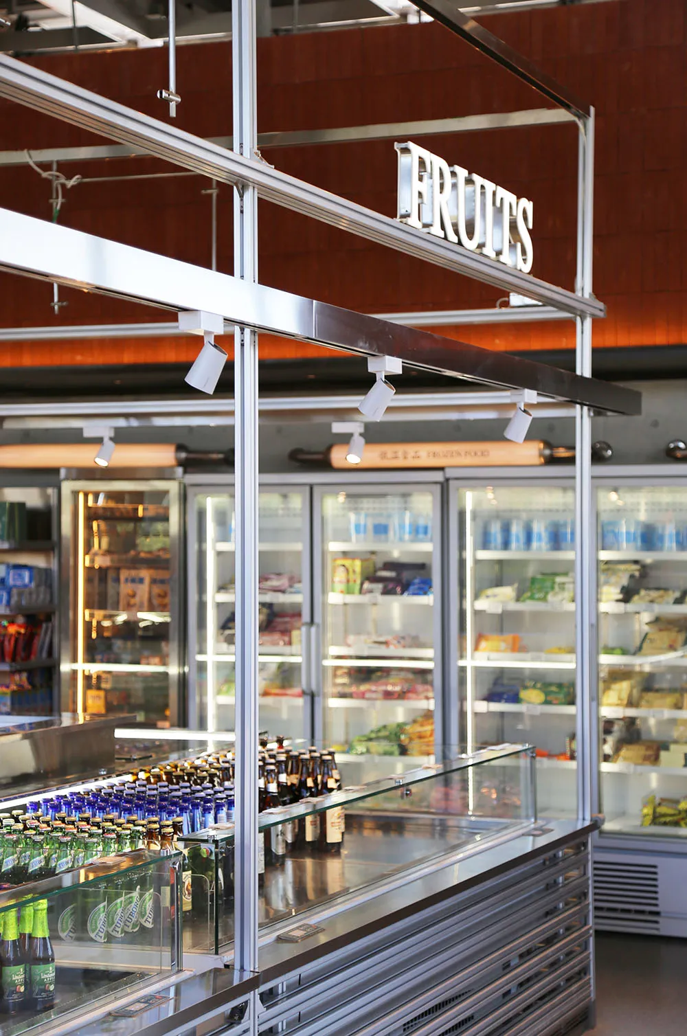 Modern supermarket interior featuring stainless steel refrigerated display cases, glass counters, and overhead metal framing with a prominent FRUITS sign.