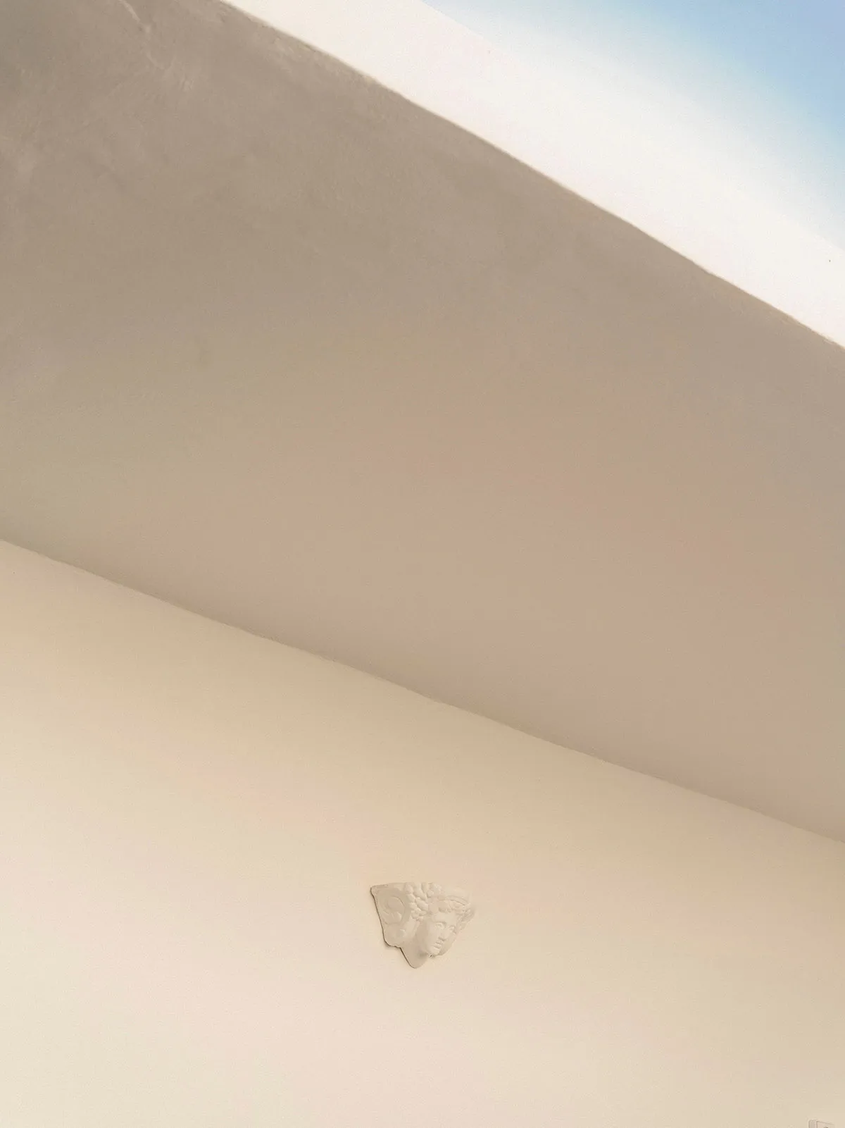 Low-angle view of a white plaster corbel with classical face motif mounted at the junction of a smooth off-white wall and ceiling under a clear blue sky.