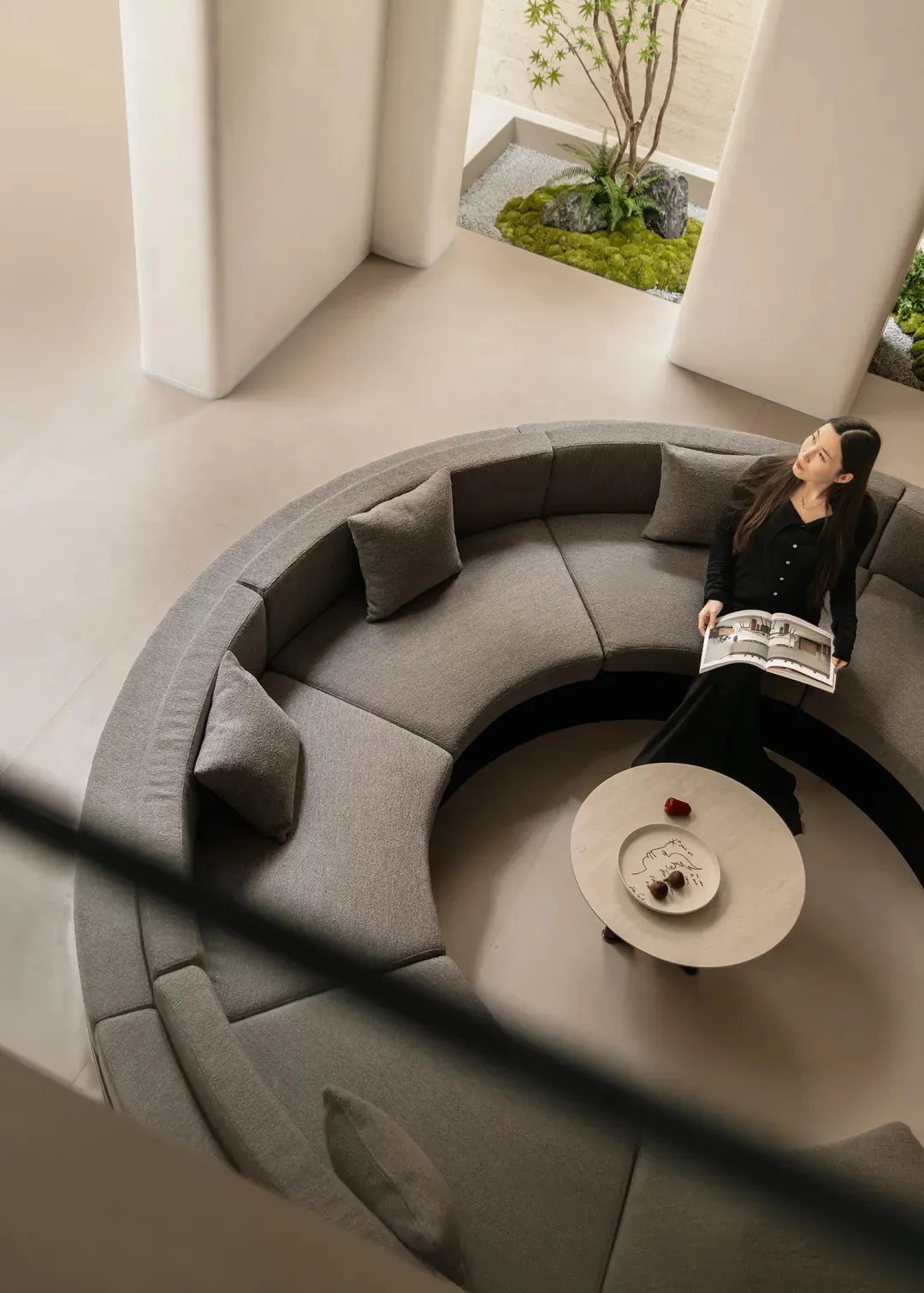 High-angle view of a modern living space featuring a large curved grey boucle sectional sofa surrounding a round coffee table, with a woman reading a book, opening to a lush sunken courtyard planted with moss, rocks and a Japanese maple tree.