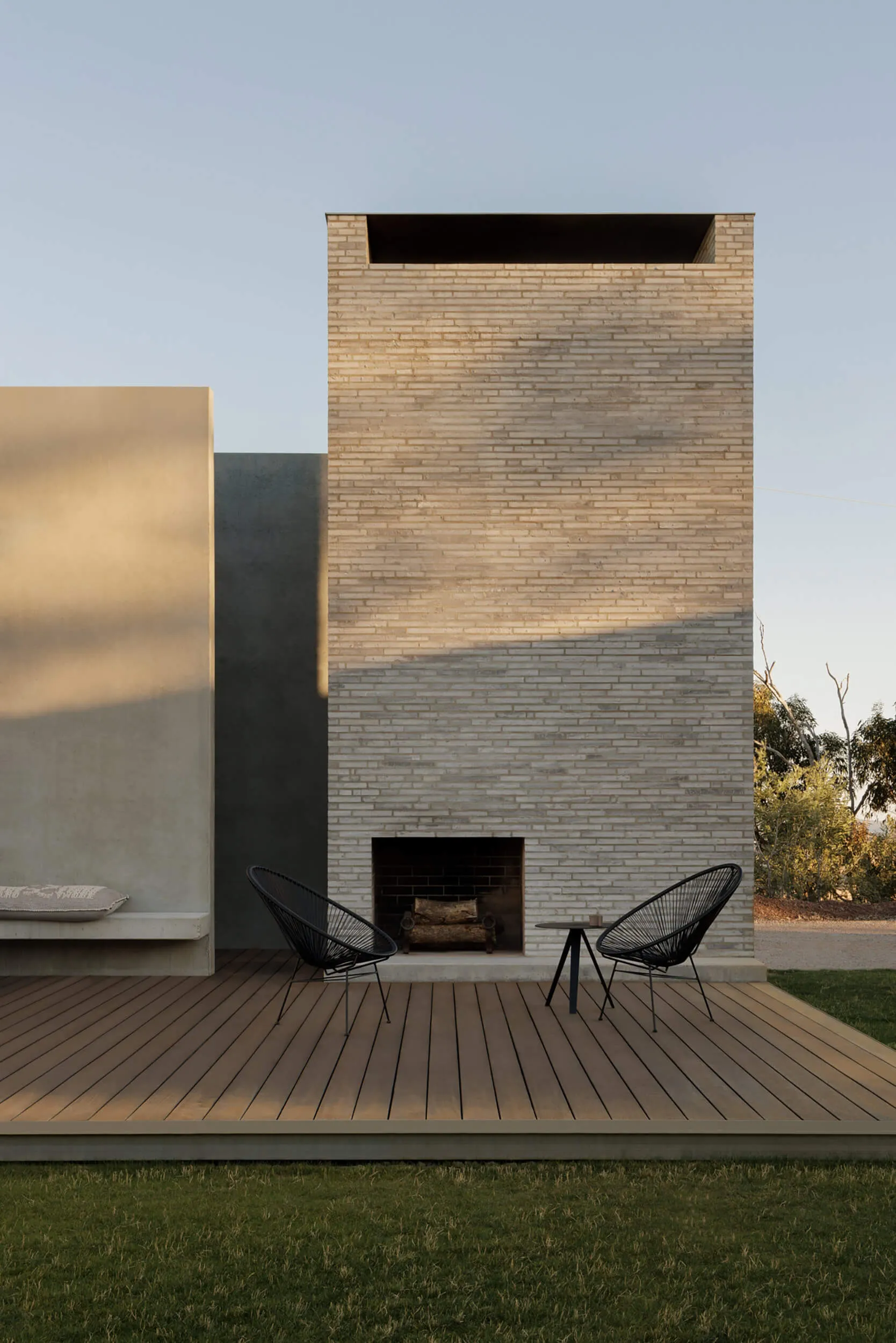 Modern outdoor deck featuring a tall light-colored brick fireplace, two black Acapulco chairs, a small side table, and a built-in concrete bench with cushion, surrounded by grass and trees.