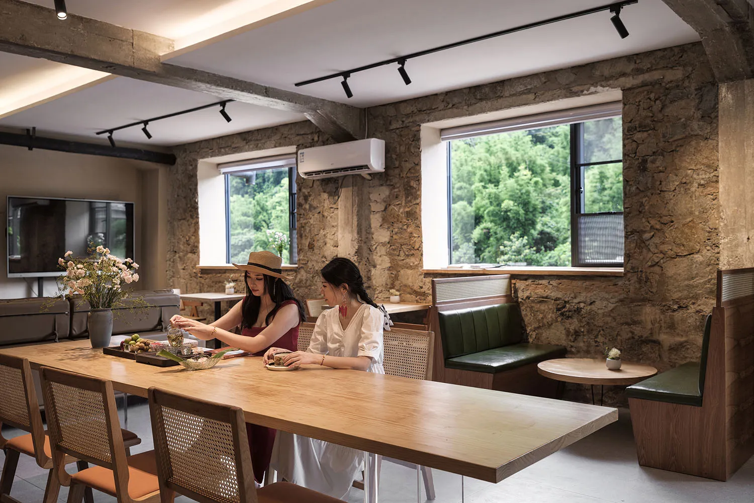 Bright modern restaurant interior featuring raw stone walls, a long solid wood communal table, rattan dining chairs, and built-in green leather banquettes with large windows overlooking a forested hillside.